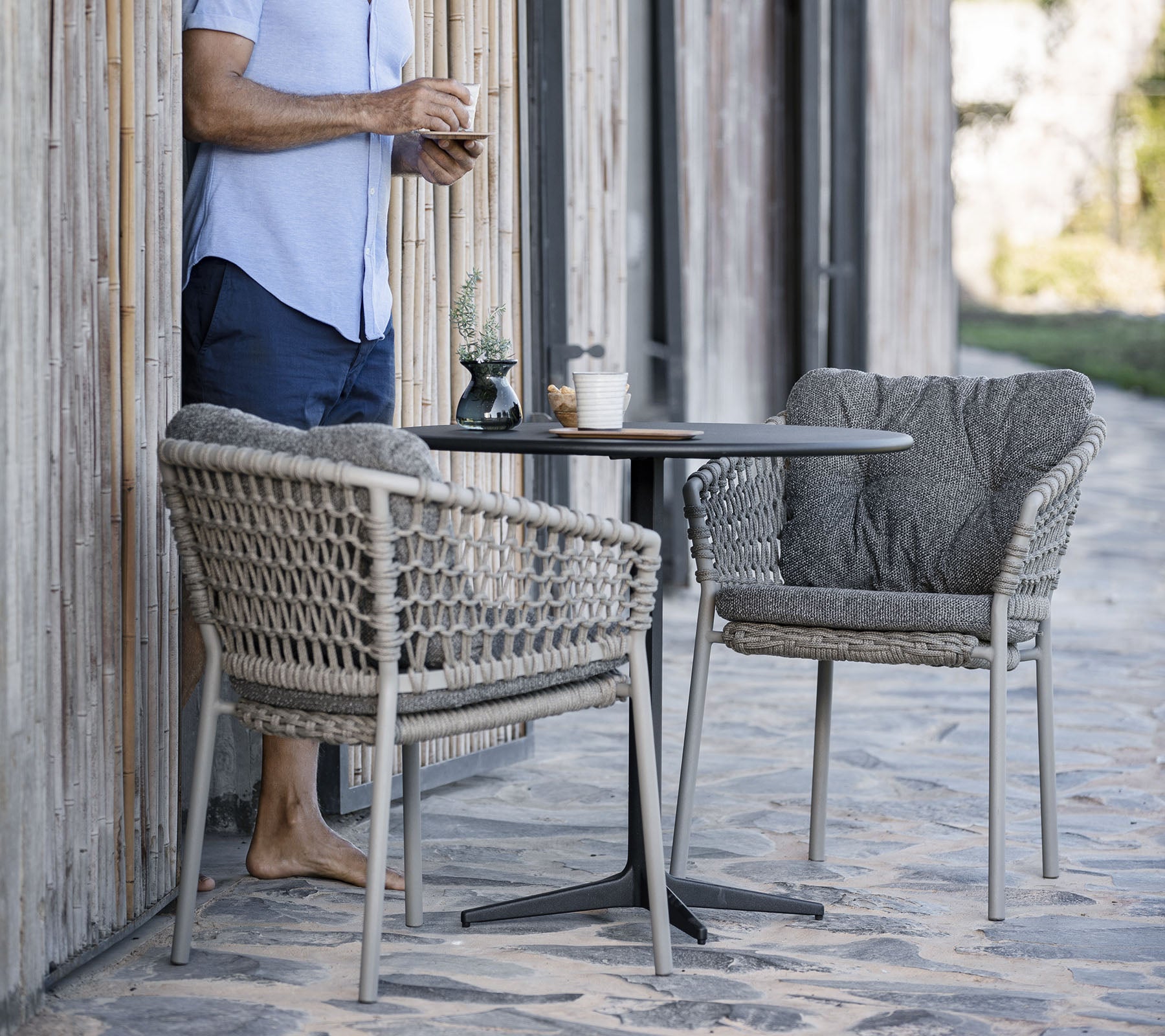 Une personne se tient à côté d'une petite table ronde avec deux chaises tressées élégantes, savourant une boisson dans un cadre détendu.