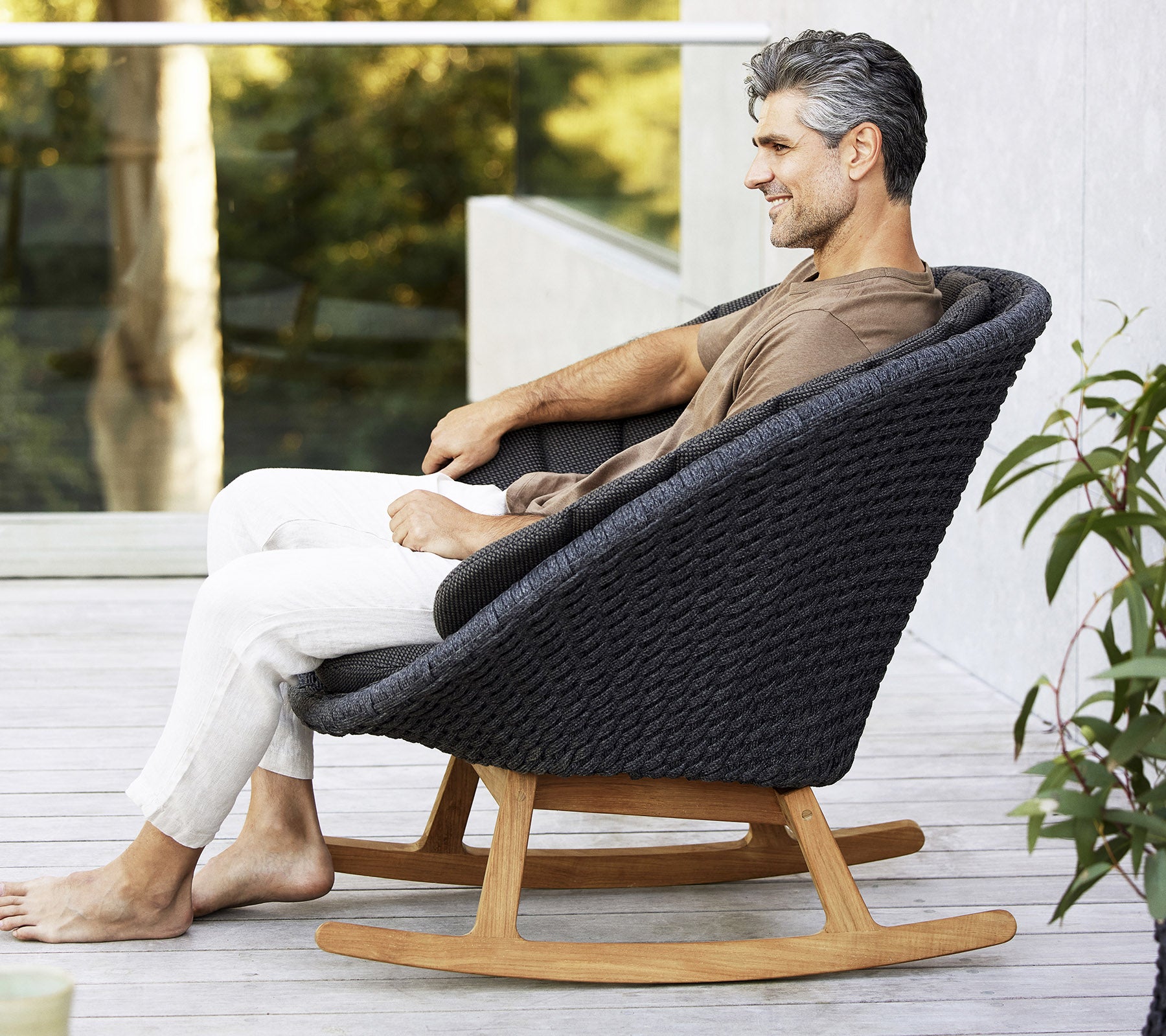 Un homme est assis, détendu, dans un fauteuil à bascule moderne et élégant, avec un design tissé et une base en bois naturel, entouré de verdure.