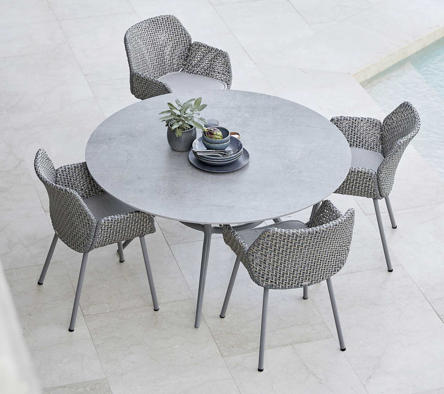 Une table ronde élégante associée à des chaises tressées, avec une plante et de la vaisselle au centre, parfaite pour les repas en plein air.