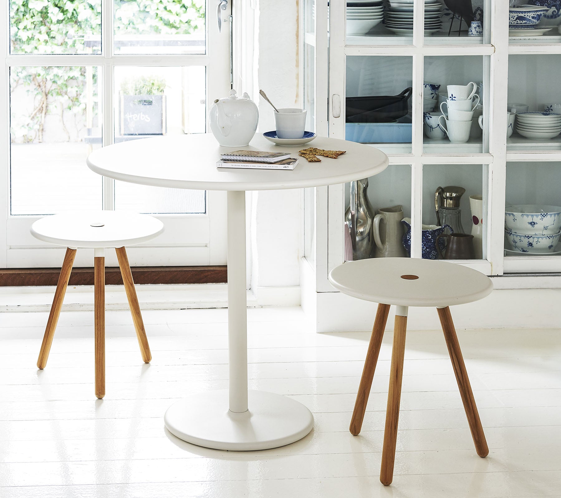 Une table ronde blanche accompagnée de deux tabourets minimalistes avec des pieds en bois, placée devant un fond d'étagères avec des ustensiles de cuisine.