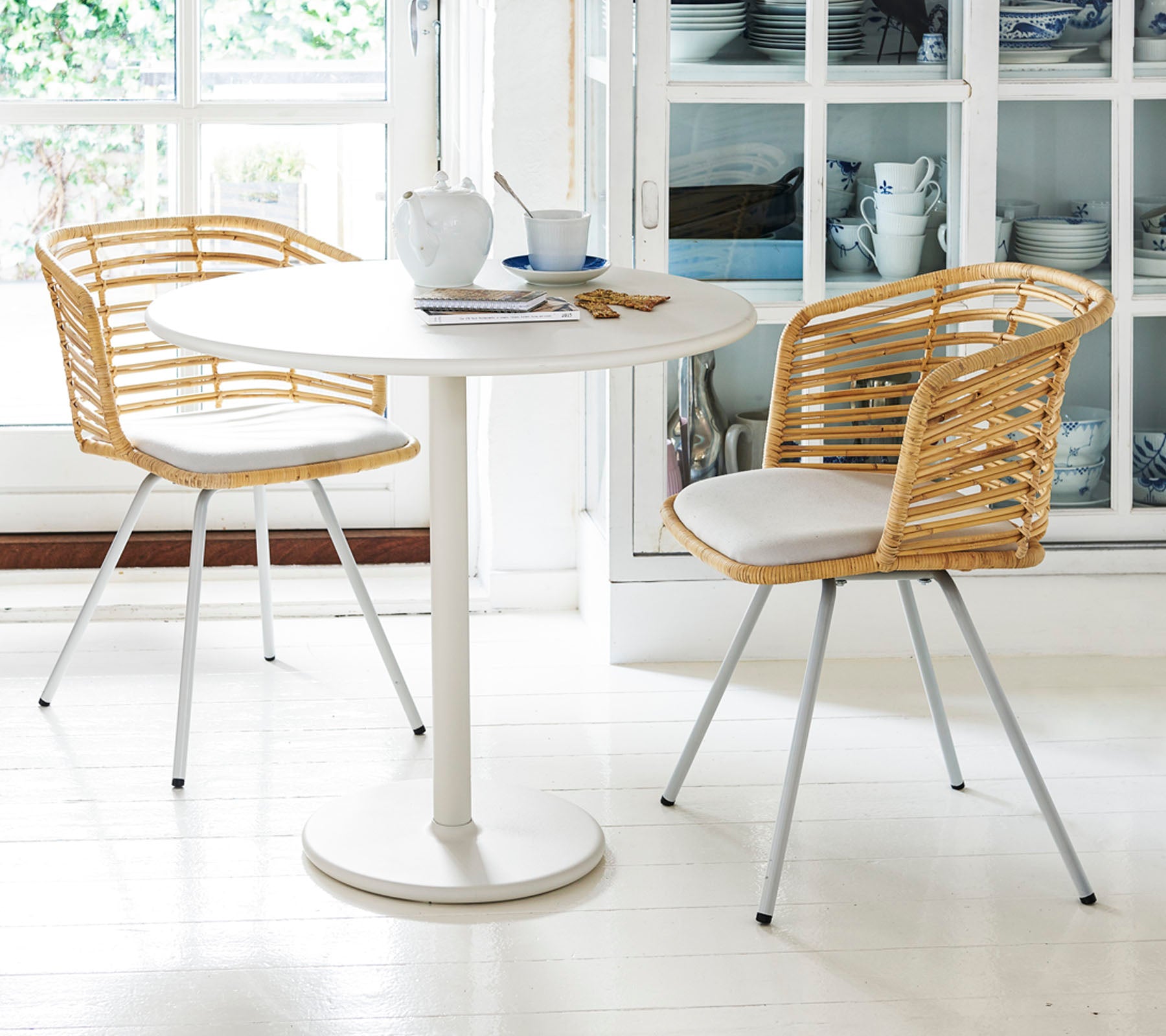 Une table ronde avec deux chaises élégantes dispose de dossiers tissés et de coussins, dressée avec des tasses à thé et une soucoupe, créant un espace repas cosy.