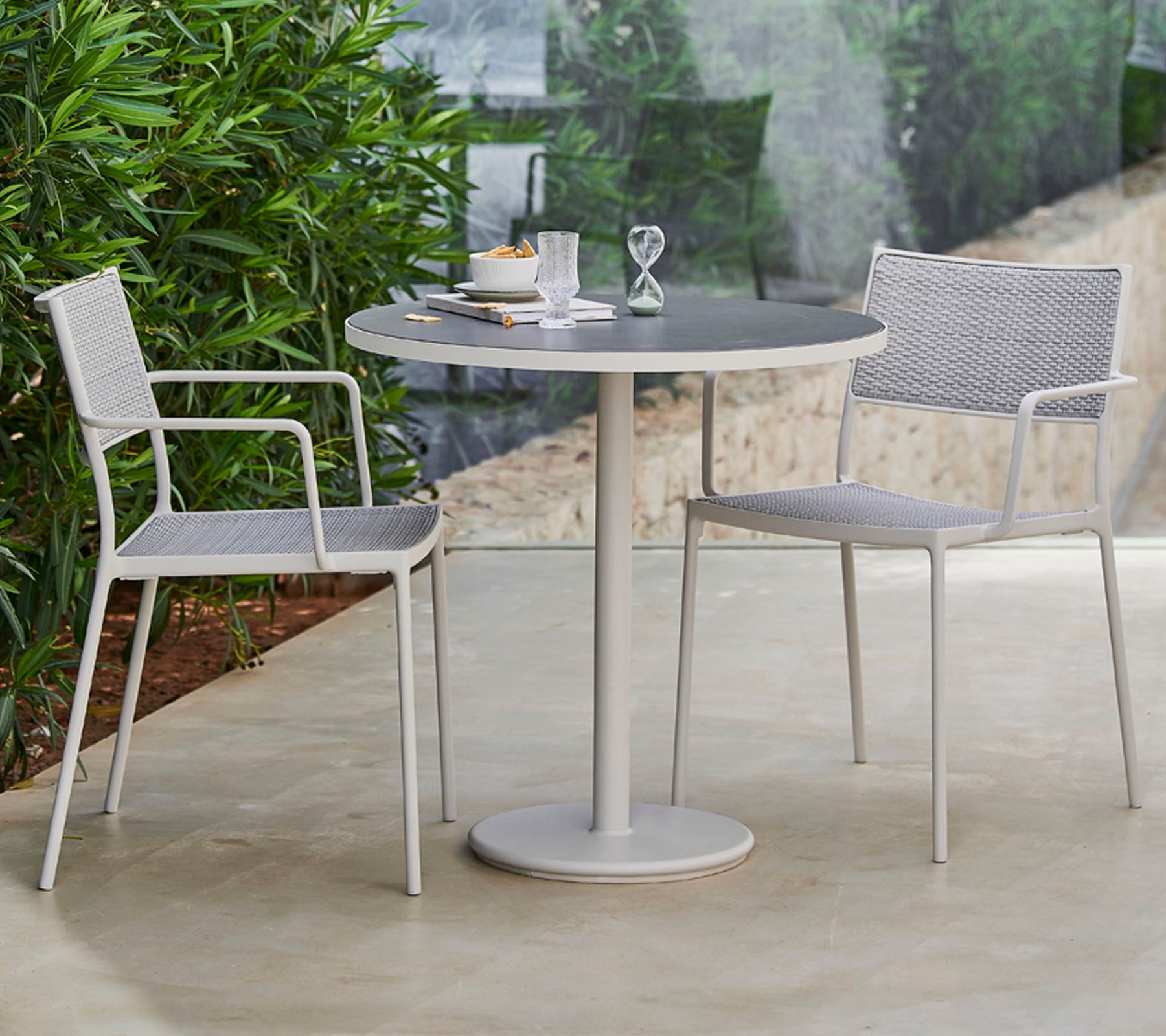 Une table ronde élégante entourée de deux chaises modernes, dressée avec de la vaisselle et un objet décoratif, encadrée par une verdure luxuriante.