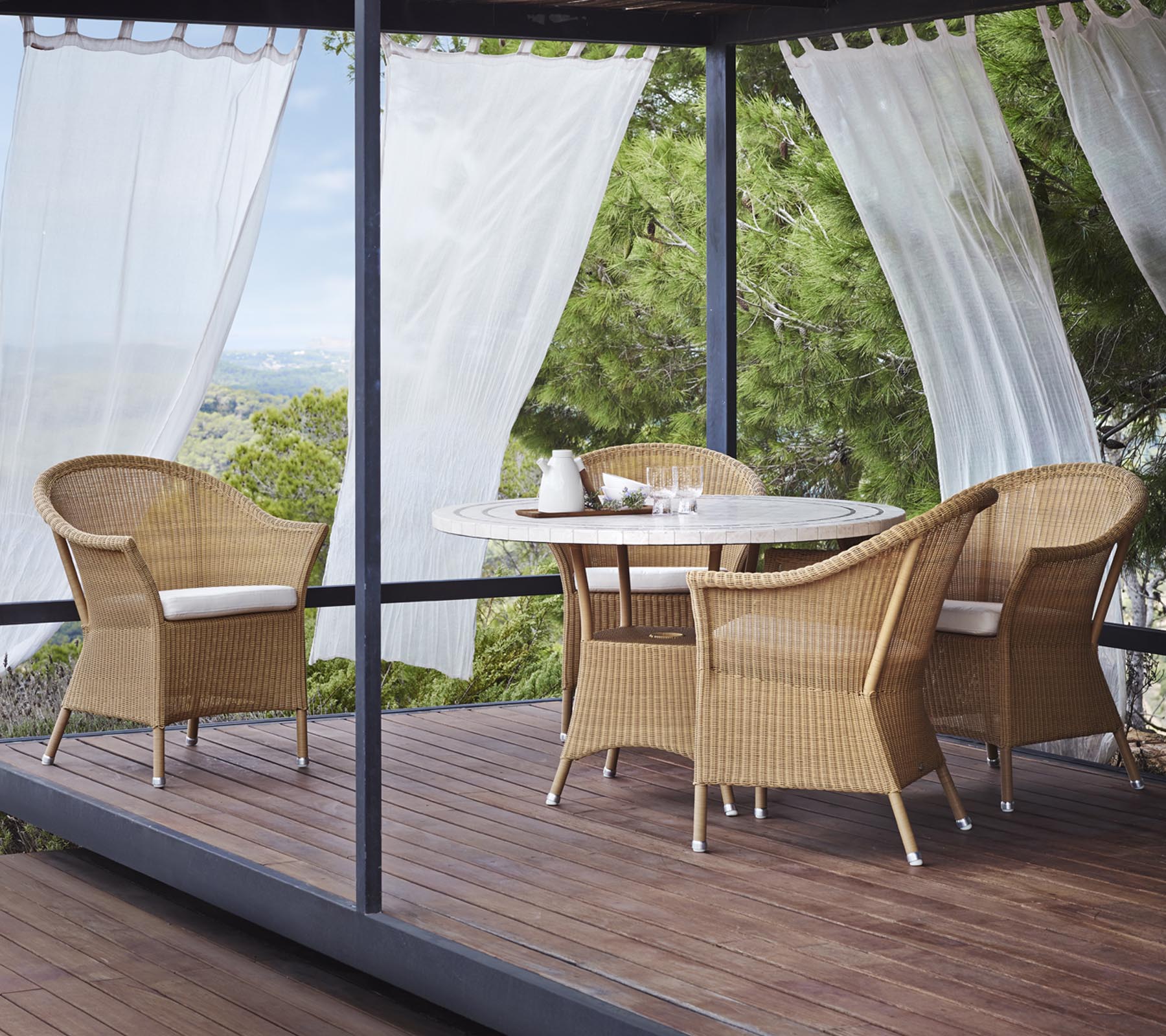 Un espace de salon élégant se compose de quatre chaises en osier et d'une table ronde, entouré de rideaux légers et de verdure naturelle.