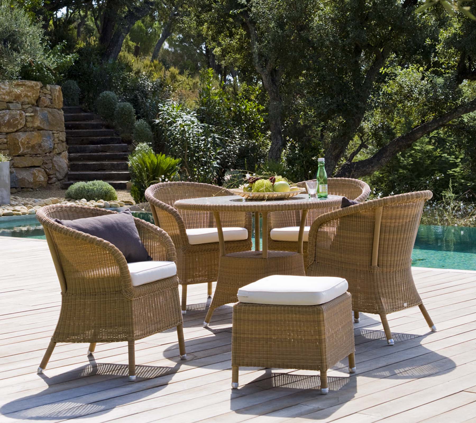 Un ensemble de mobilier d'extérieur élégant comprenant une table ronde, quatre chaises tressées et un pouf, entouré d'une végétation luxuriante et d'un cadre paisible.