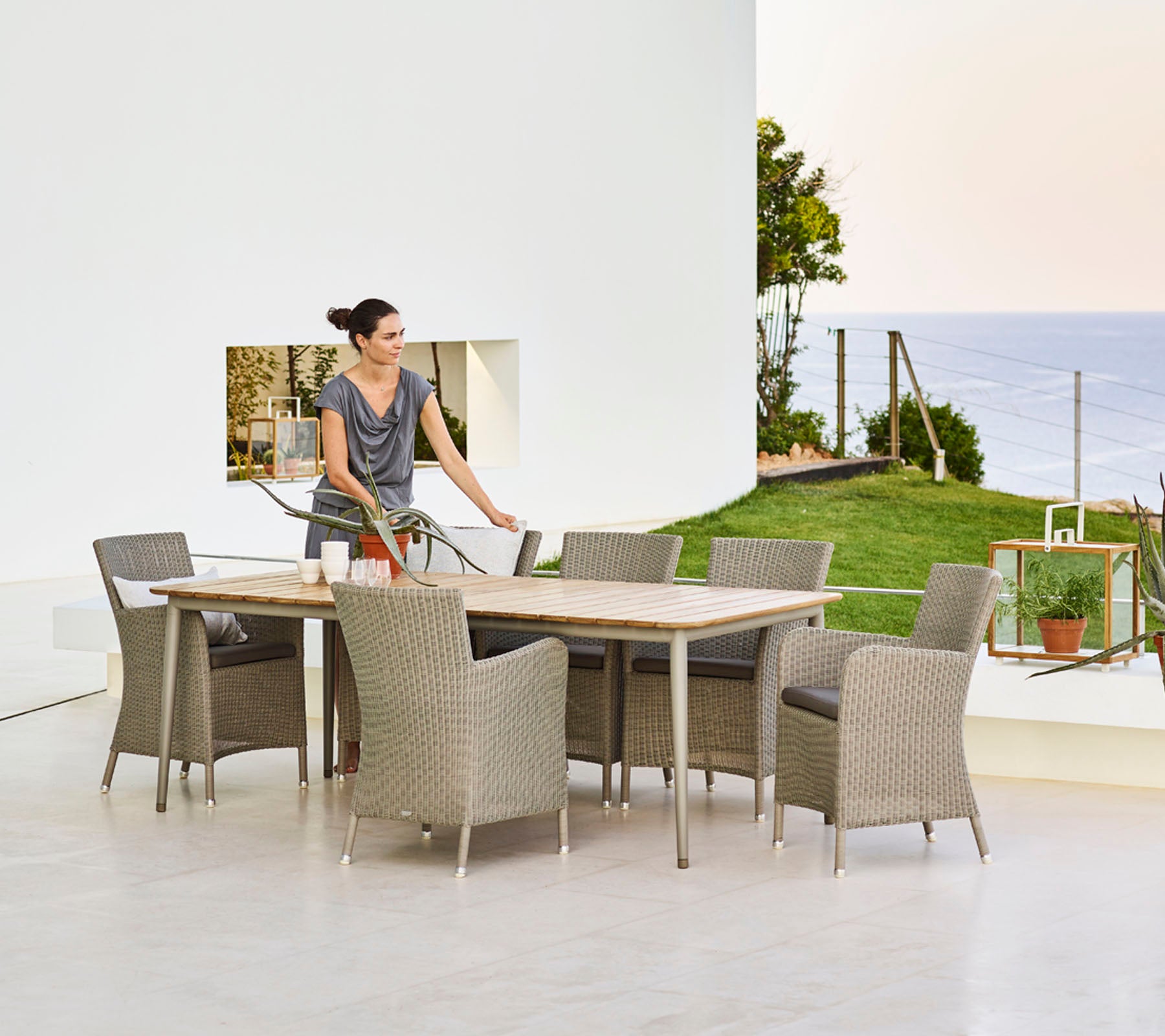 Une femme met la table à manger, entourée de chaises en osier, surplombant une vue pittoresque avec de la verdure et de l'eau en arrière-plan.