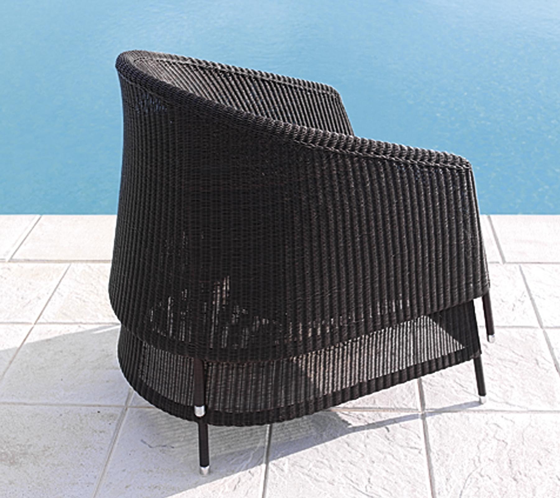 Chaise en rotin noire courbée avec un design épuré, positionnée près d'une piscine sur un sol carrelé blanc.