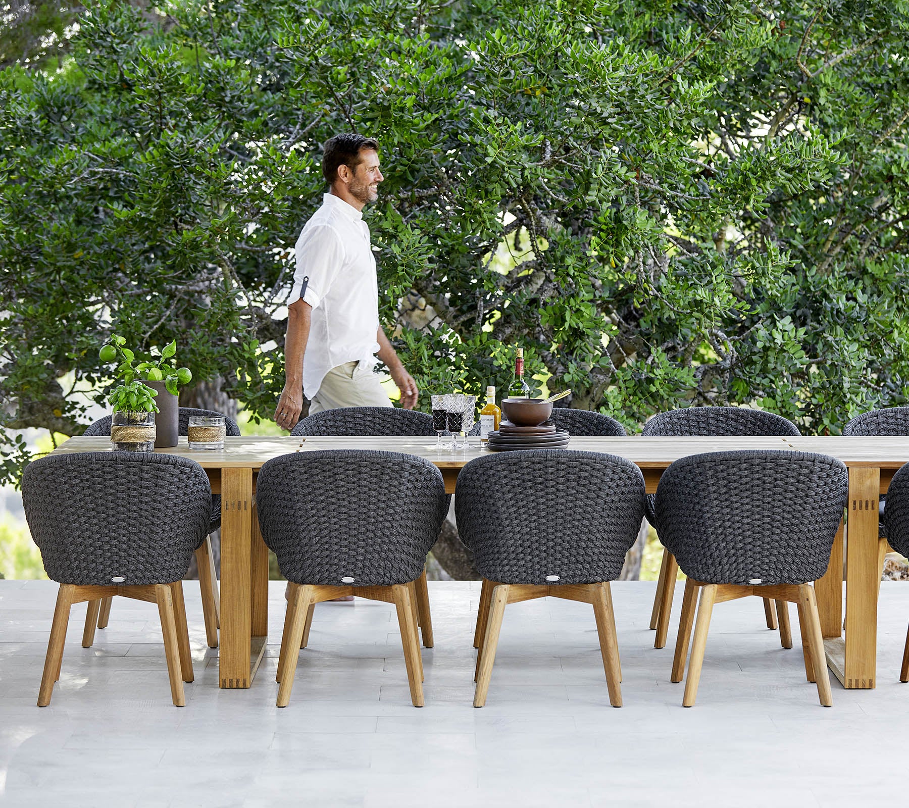 Un homme passe devant une grande table à manger en bois entourée de chaises modernes en osier et d'une verdure en arrière-plan.