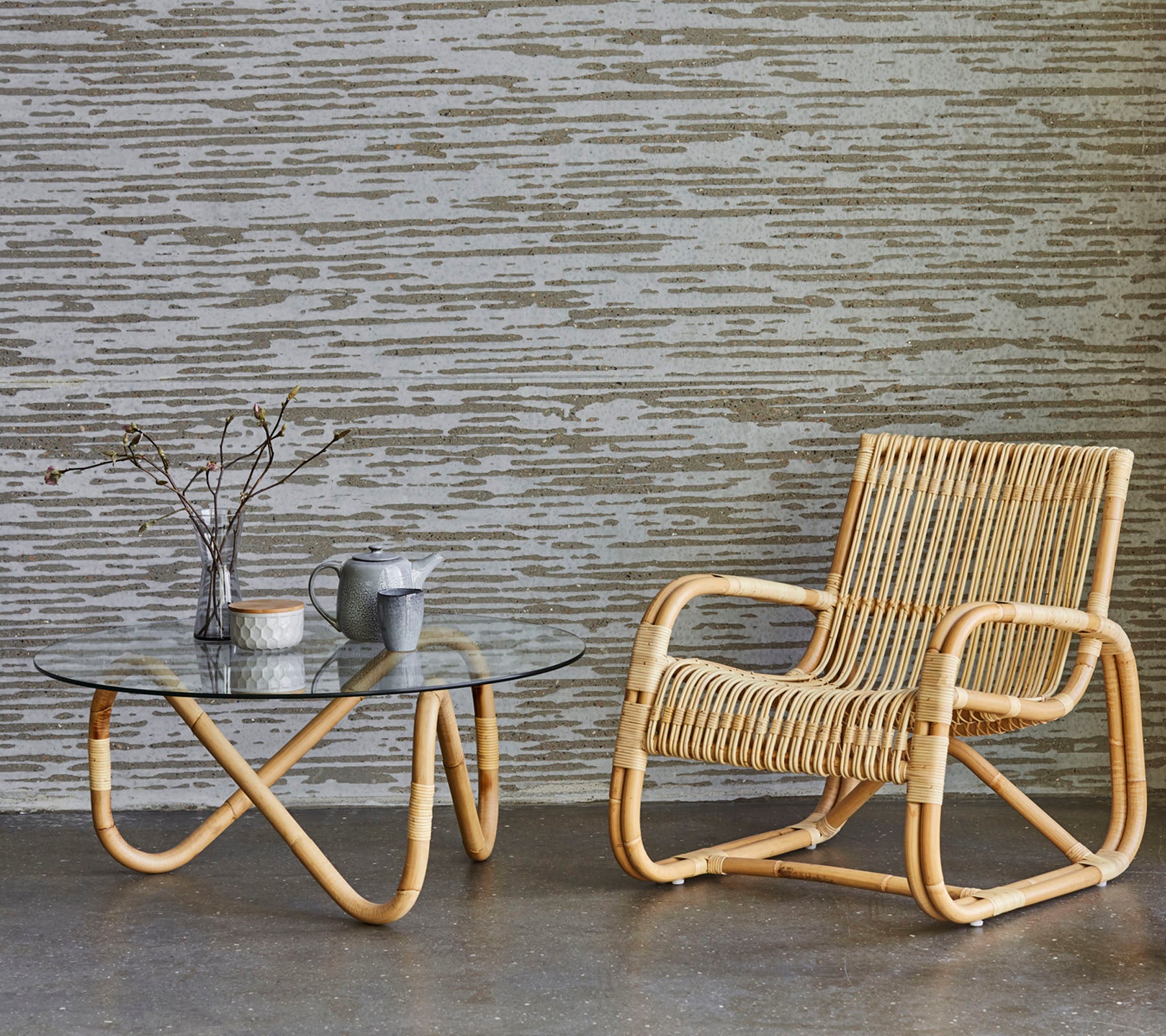 Une chaise en rotin élégante à côté d'une table basse en verre, avec un vase contenant des branches et une petite théière, sur un fond texturé.