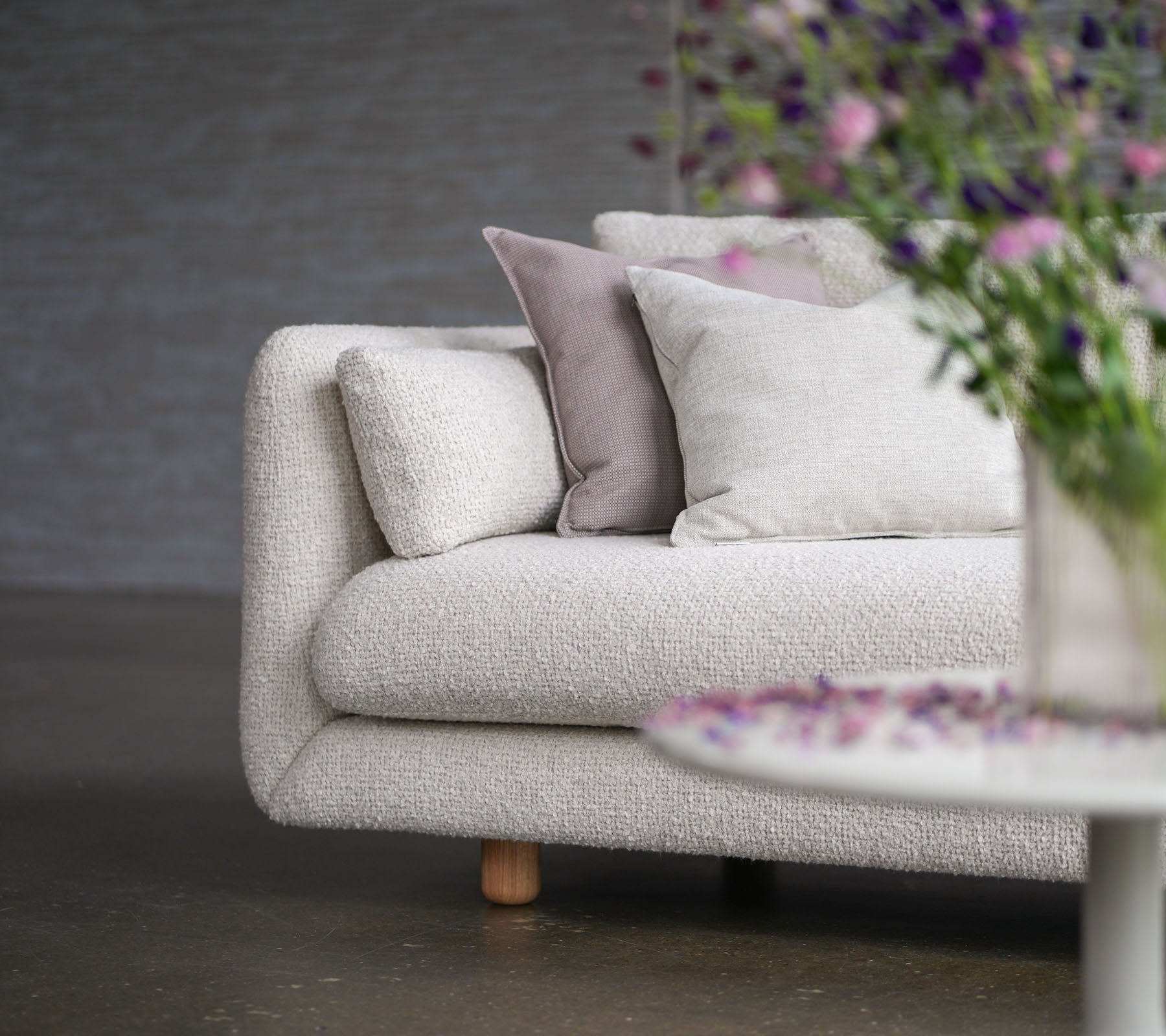 Canapé doux avec un revêtement couleur sable, des coussins décoratifs, des pieds en teck et un vase de fleurs, mettant en valeur le style et l'élégance de Cane-line.
