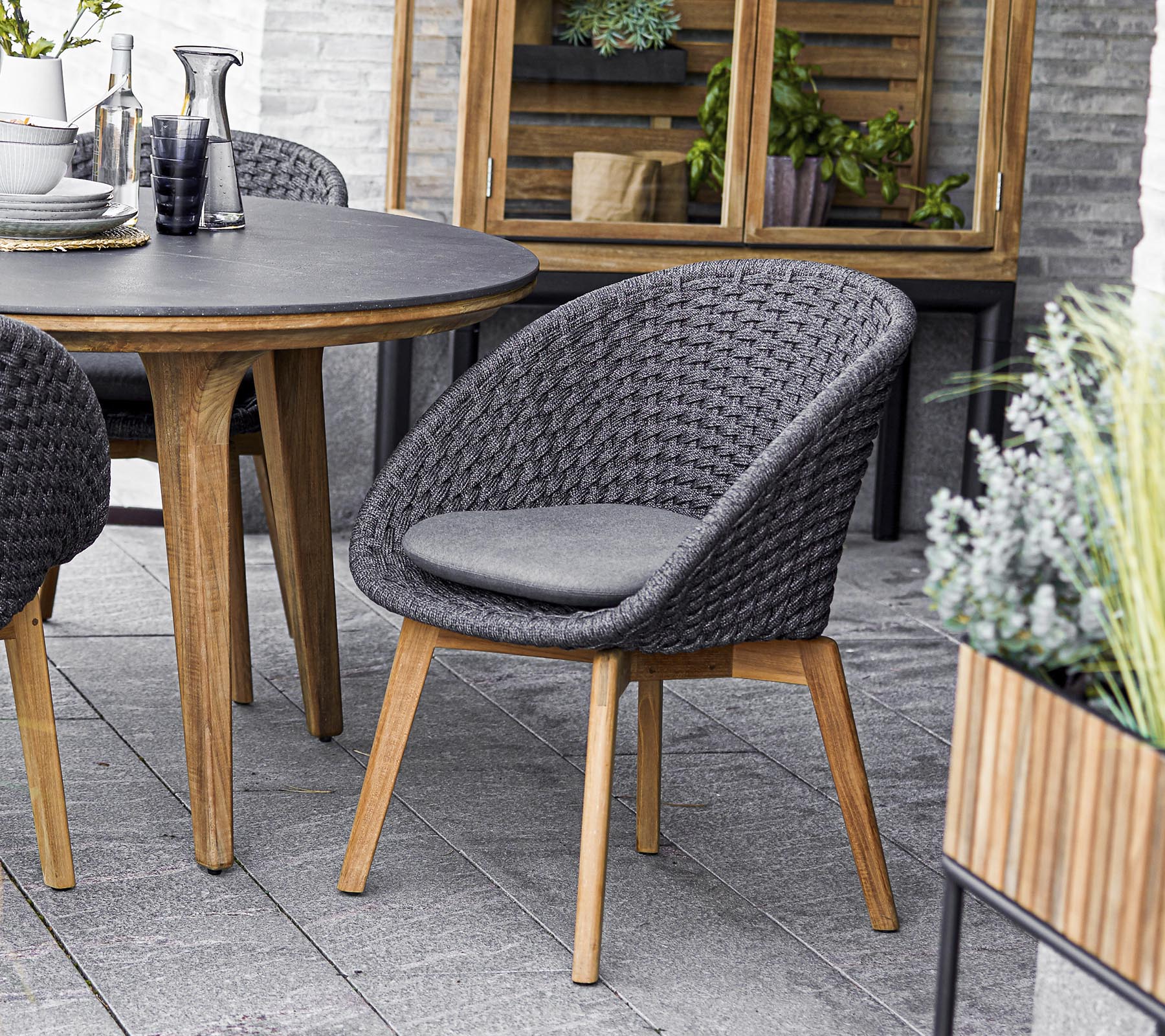 Chaise tissée élégante avec un siège rembourré, accompagnée d'une table ronde et entourée de verdure dans des jardinières. Parfait pour des rassemblements sociaux.