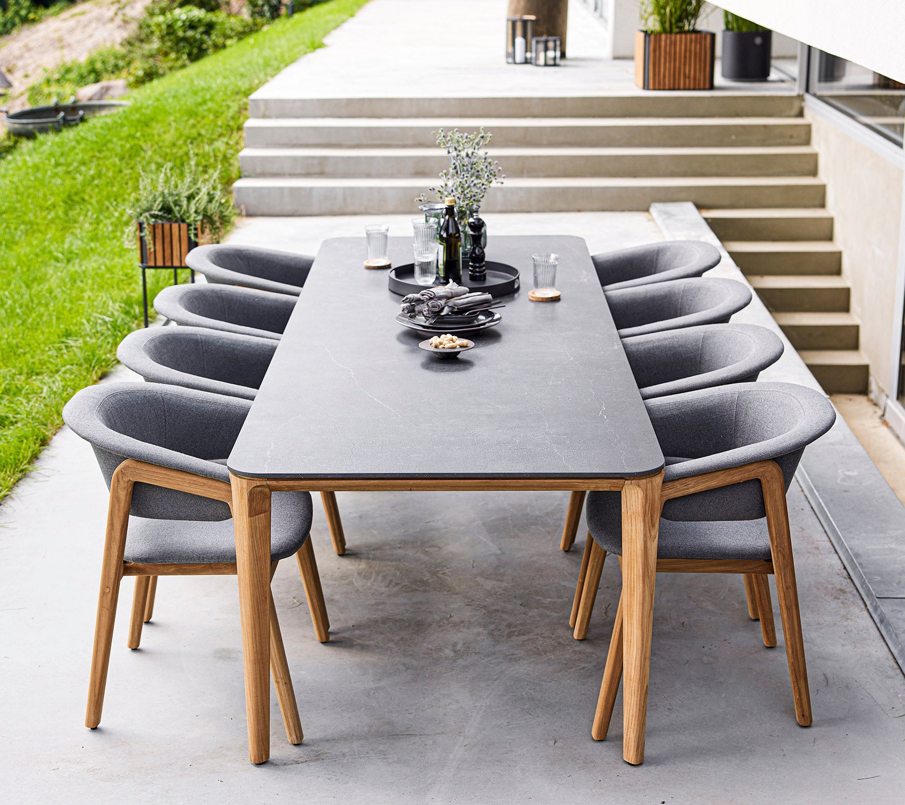Une table à manger moderne avec huit chaises grises, présentant des objets décoratifs et des plantes sur la table, située dans un espace extérieur élégant.