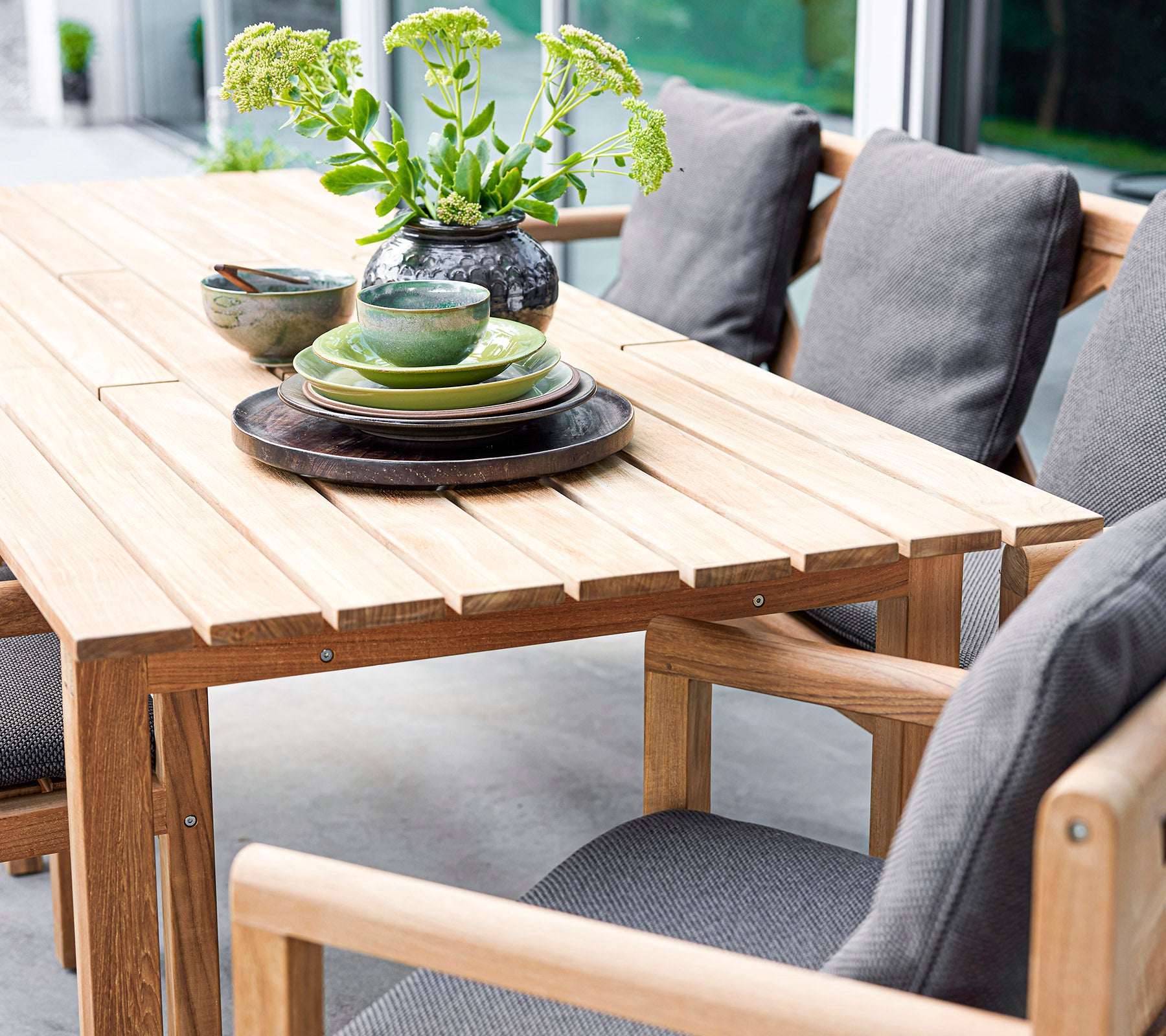 Ensemble de table en bois élégant avec des coussins, des assiettes et des bols, accentué par un vase avec du feuillage. Parfait pour des rassemblements accueillants.