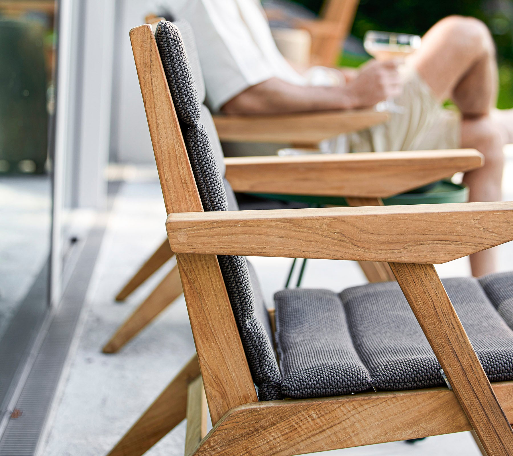 Une chaise en bois avec un coussin gris au premier plan, avec une personne se détendant à proximité tenant une boisson, capturant un moment de loisirs.
