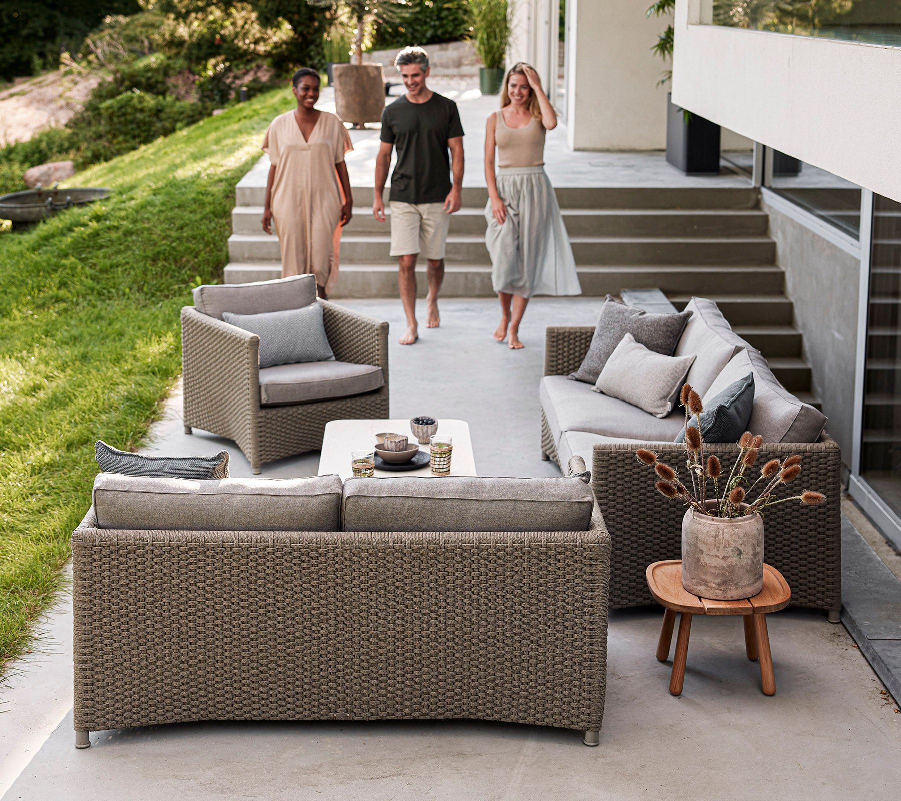 Trois personnes marchent vers un espace extérieur élégant avec un mobilier tissé confortable, des coussins décoratifs et une table basse.