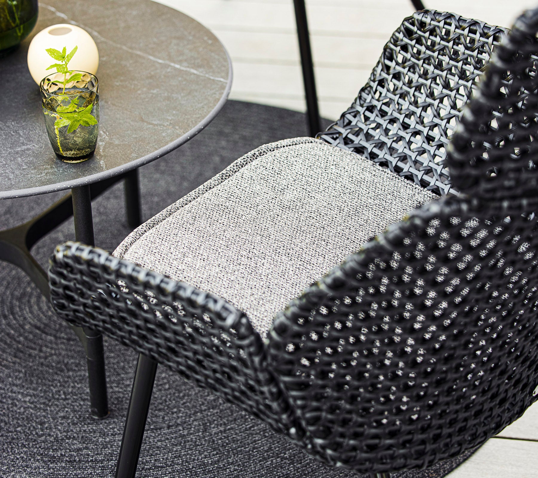 Un gros plan d'une chaise tissée élégante et d'une petite table avec un verre décoratif et une plante, assortie d'un tapis circulaire confortable.
