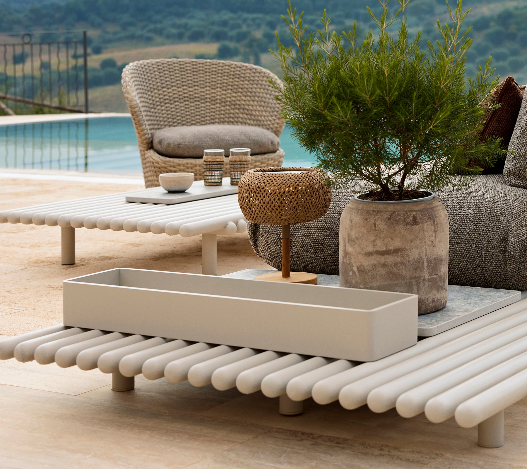 Boîte de rangement en aluminium sur une table à lattes, entourée de mobilier de jardin et de verdure, avec une vue pittoresque en arrière-plan.