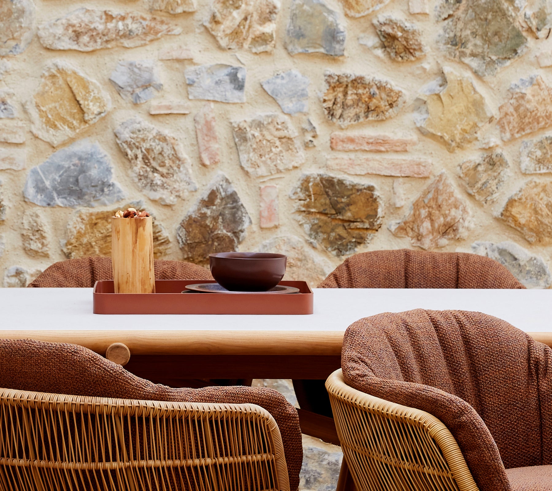 Un aménagement cosy avec un plateau de club rouge désert, un bol marron et des chaises tressées élégantes devant un mur en pierre.