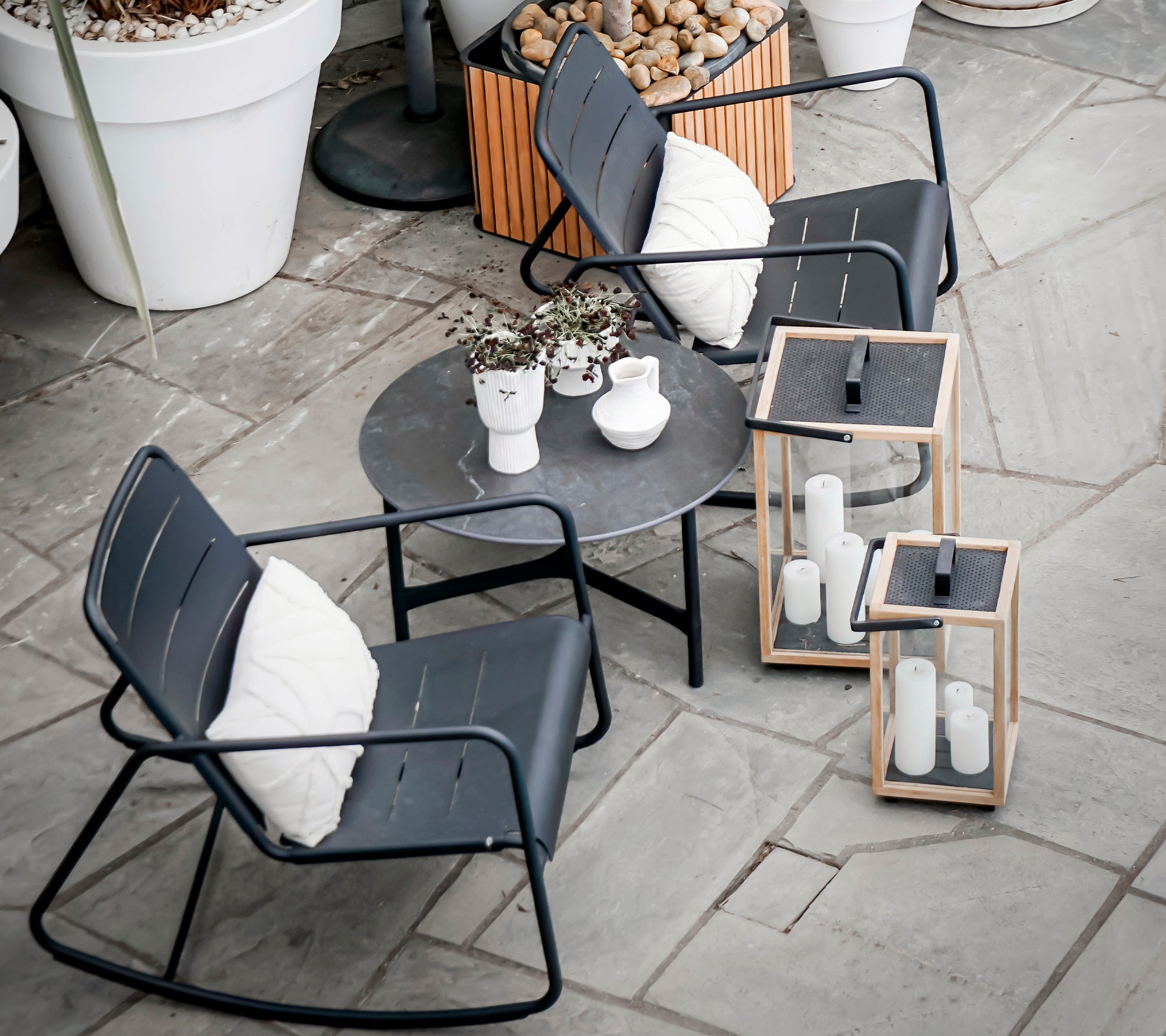 Deux chaises à bascule modernes et une table ronde avec des plantes décoratives et des lanternes, placées sur une surface en pierre texturée.