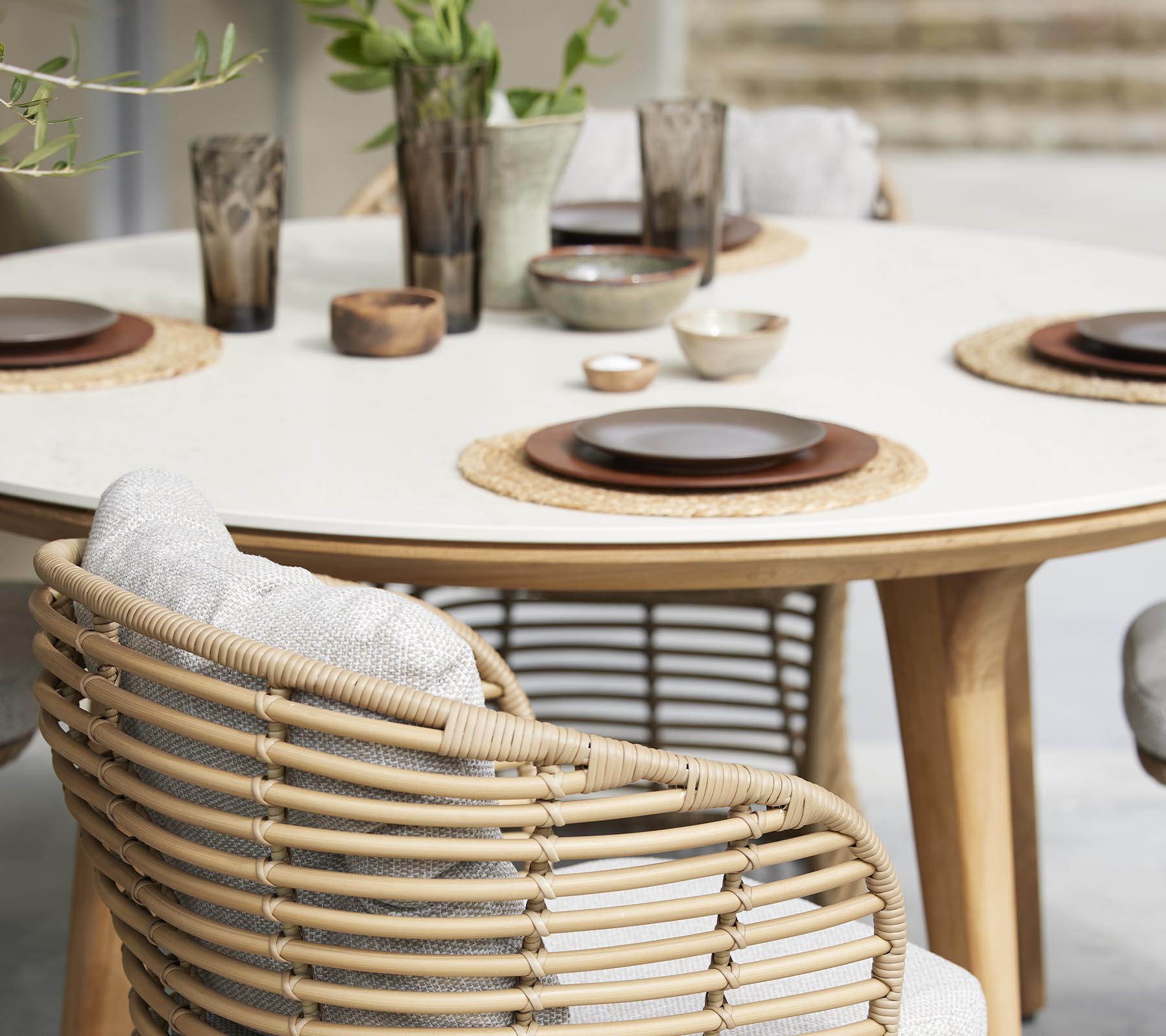 Une table à manger élégante dressée avec des assiettes, des bols et des verres décoratifs, entourée de chaises tressées et d'éléments naturels.