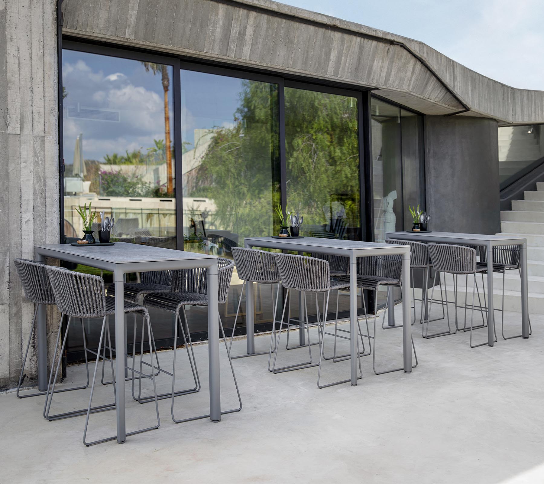 Espace de seating moderne avec des tables et des chaises grises, de grandes fenêtres reflétant la verdure à l'extérieur. Esthétique de design minimaliste.