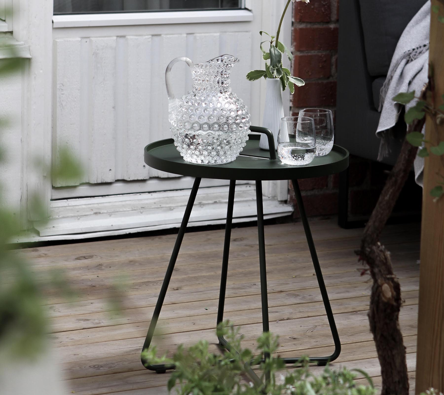 Une table d'appoint verte élégante avec un pichet en cristal et deux verres, complétée par une verdure fraîche dans la zone environnante.
