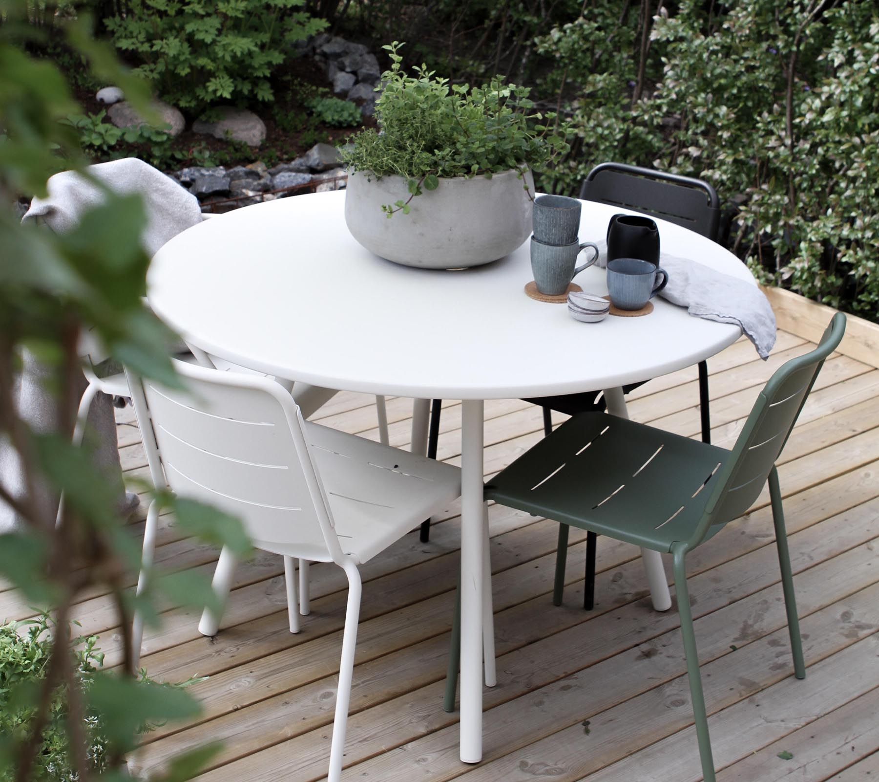 Une table ronde entourée de quatre chaises, avec une plante en pot et des mugs à café sur le plateau. Une verdure luxuriante en arrière-plan.