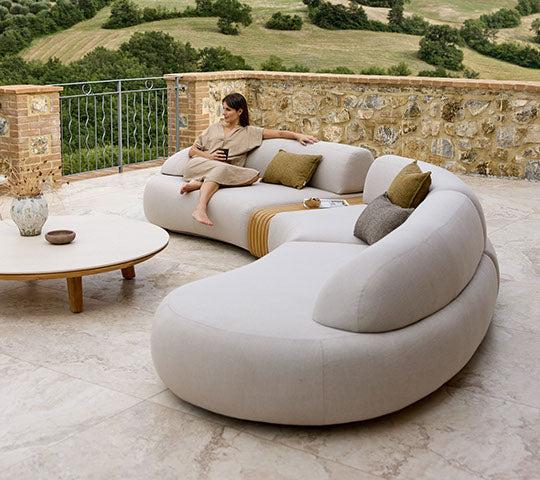 Un canapé courbé et moelleux avec des coussins et une table basse en bois, accompagné d'une personne assise confortablement en profitant de la vue.