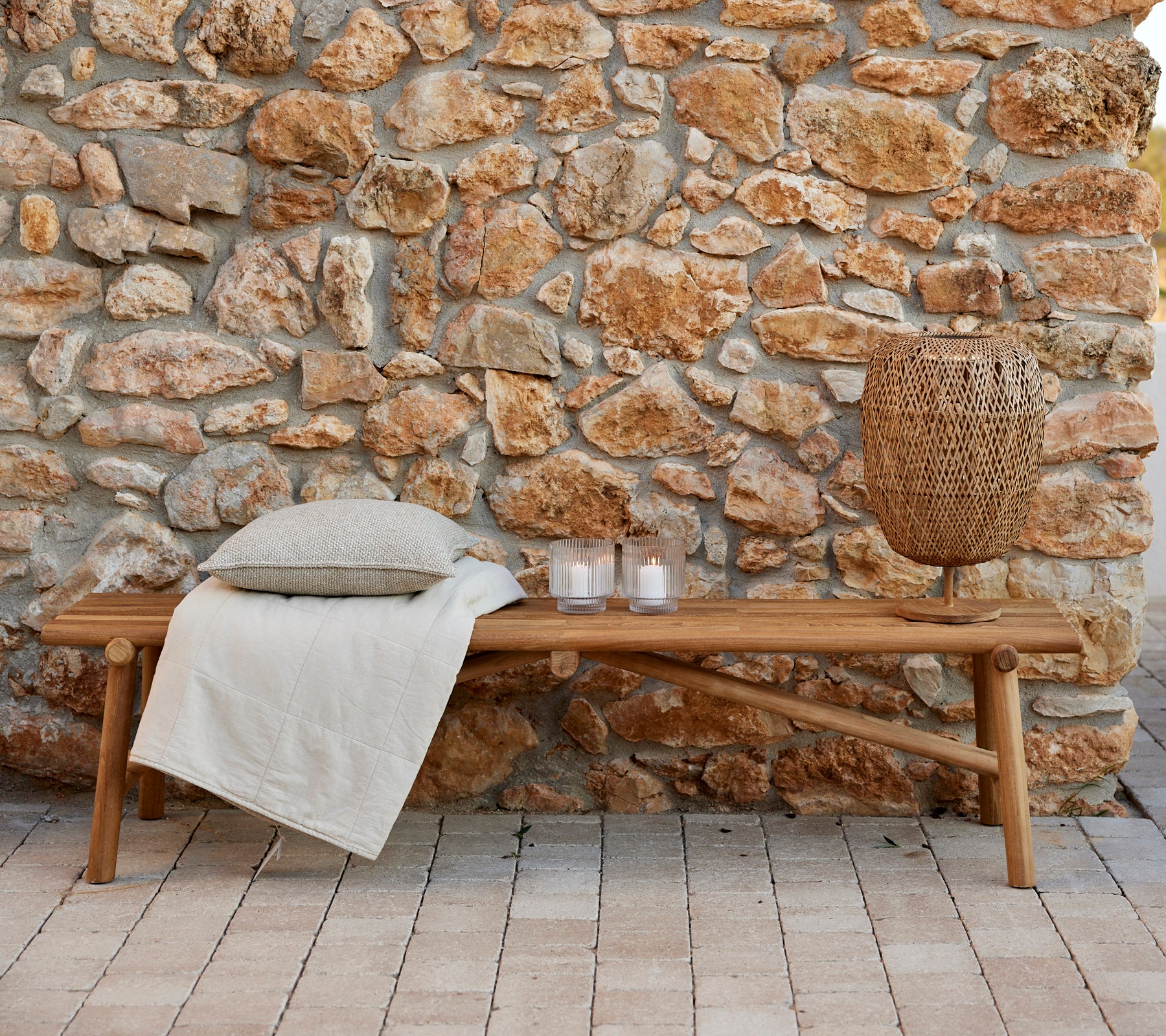 Banc en teck avec une couverture confortable, des coussins décoratifs, des bougies et une lampe tissée, contre un mur en pierre naturelle. Idéal pour se détendre.