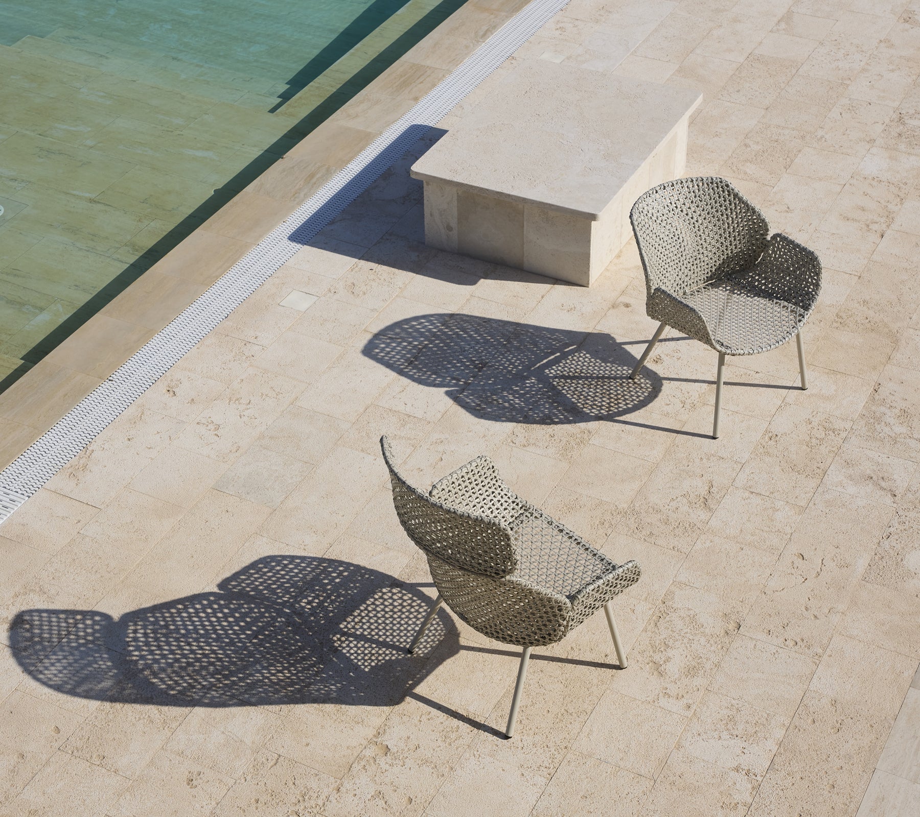 Deux chaises longues élégantes avec des designs uniques projetant des ombres, placées près d'une surface d'eau calme et d'une table en pierre.