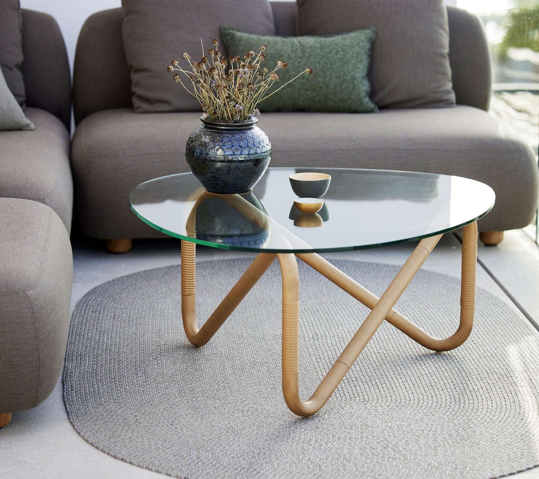 Table basse ondulée avec un plateau en verre et une base en bois, accentuée par un vase et un petit bol, entourée de sièges doux et de couleur neutre.