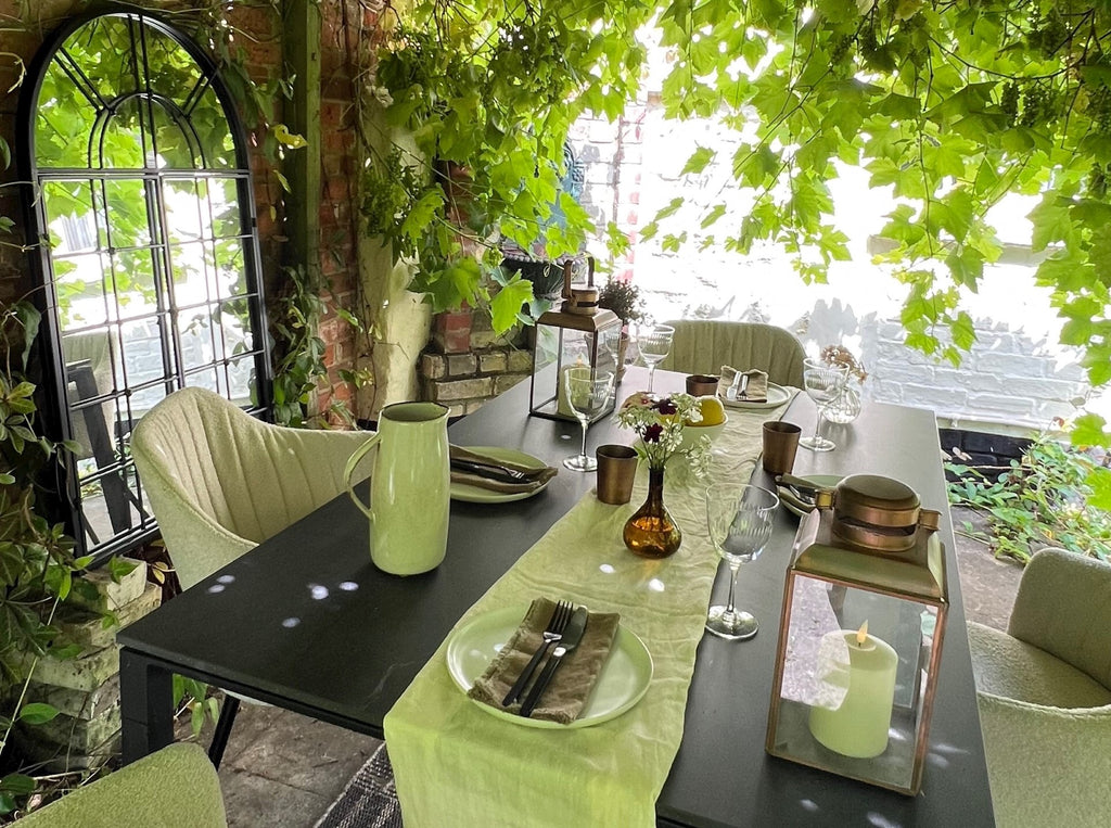 Pergola with green lush grapevines, sand bouclé outdoor chairs, ceramic black outdoor table, white table runner, glass, dishes 