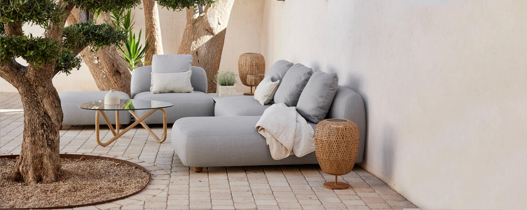 Espace salon élégant avec de confortables canapés gris, des coussins décoratifs, un plaid douillet et des éléments naturels comme un arbre en pot et des tables d'appoint tressées.