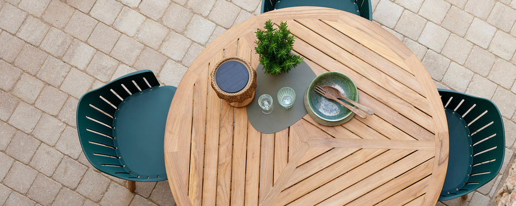 A round wooden table with a small plant, two plates, and cutlery, accompanied by two green chairs.