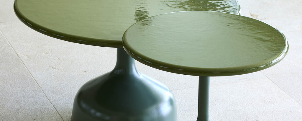 Two modern tables with a glossy glaze finish, featuring a unique design with varying heights and round tops in a muted green color.