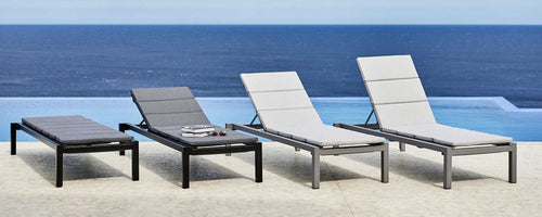 Four modern sunbeds in a serene setting by the water, featuring sleek designs in contrasting colors, perfect for relaxation.