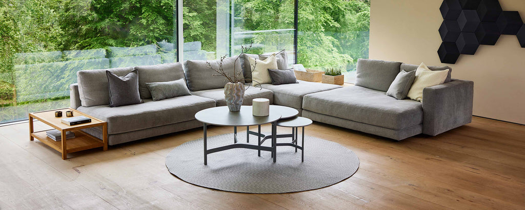 Un salon spacieux et moderne comportant un grand canapé d'angle gris, un tapis rond et des tables basse élégantes, avec de la verdure visible à l'extérieur.
