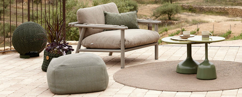 Espace salon élégant avec une chaise confortable, une table ronde, un pouf et des accessoires décoratifs disposés sur un tapis texturé.