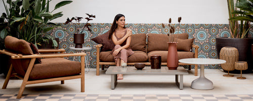 Un coin salon élégant avec un canapé brun, un fauteuil et une table décorative, entouré d'une végétation luxuriante et d'une décoration à motifs.