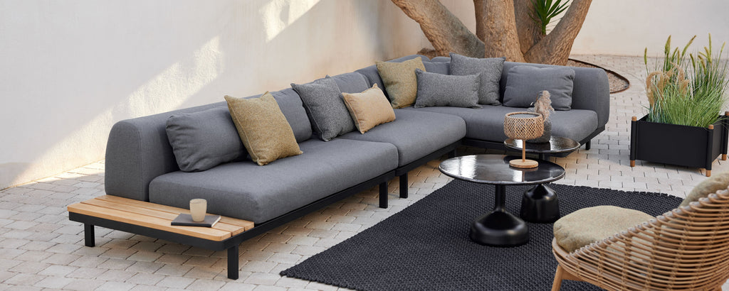 Espace lounge avec un canapé sectionnel moderne avec des coussins décoratifs, une table basse ronde et un tapis élégant, complété par des plantes.
