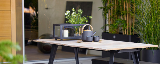 Table moderne avec lanterne noire, théière et tasse entourées de verdure.