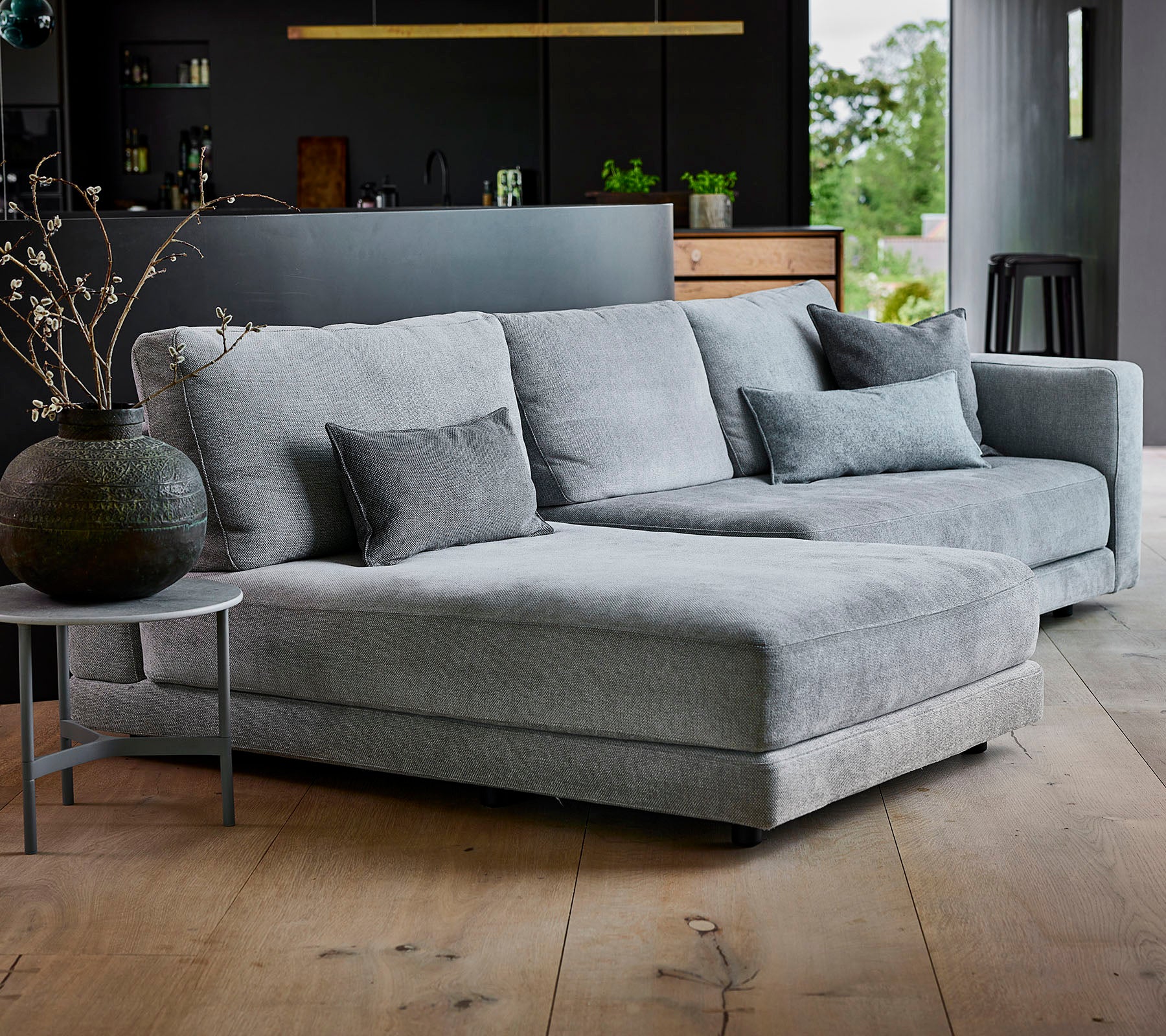 Canapé sectionnel gris avec tissu texturé et coussins décoratifs, accompagné d'une table d'appoint ronde et d'un vase élégant.