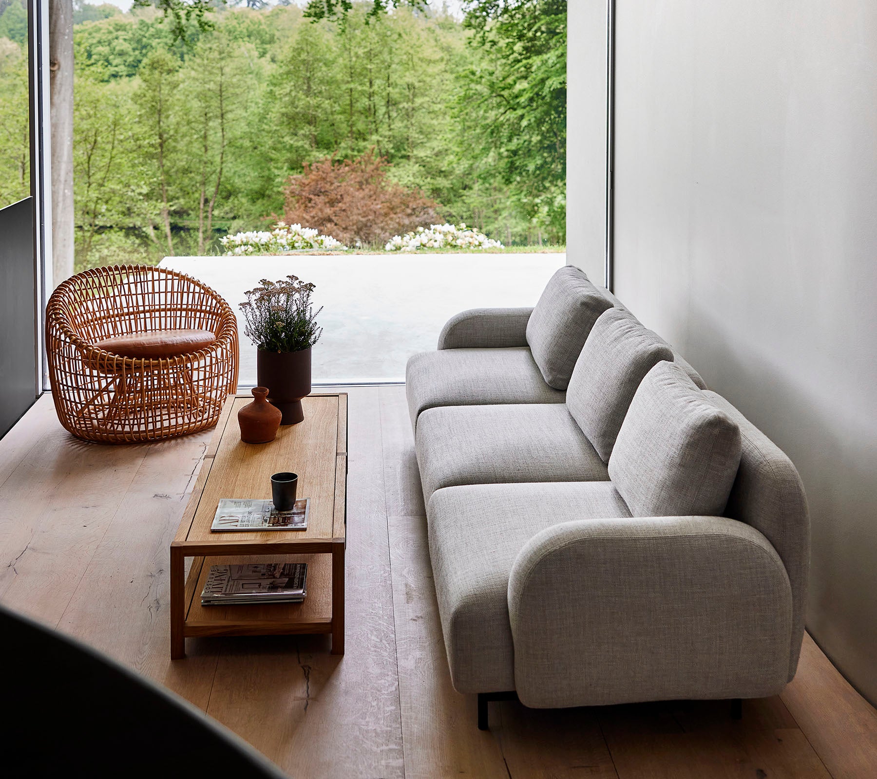 Canapé moderne en tissu clair associé à une table basse en bois et une chaise en osier, entouré de verdure. La lumière naturelle enhance l'ambiance sereine.