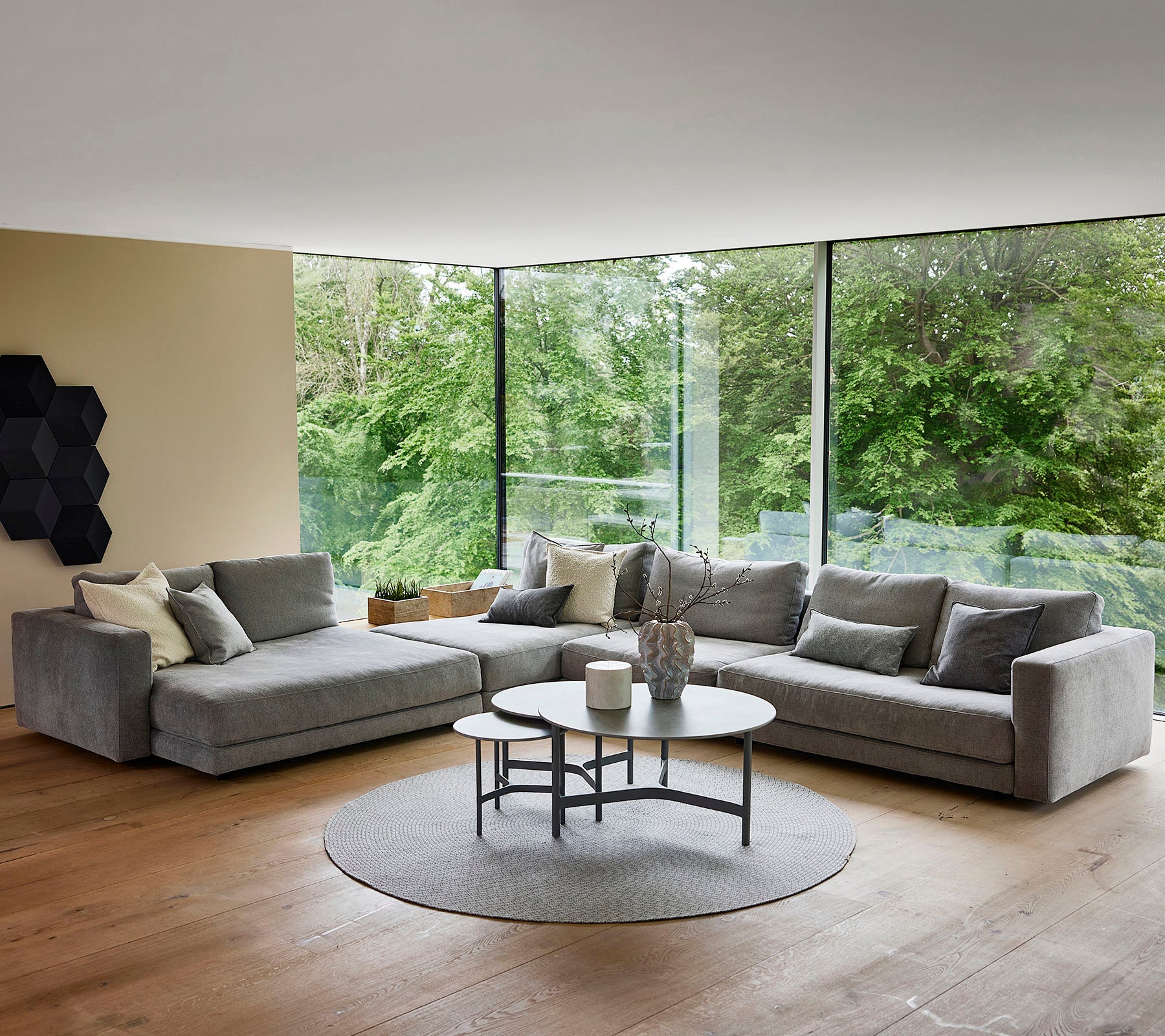 Un espace de vie moderne avec un canapé d'angle gris orné de coussins décoratifs, une table basse ronde et un tapis circulaire, entouré de verdure.