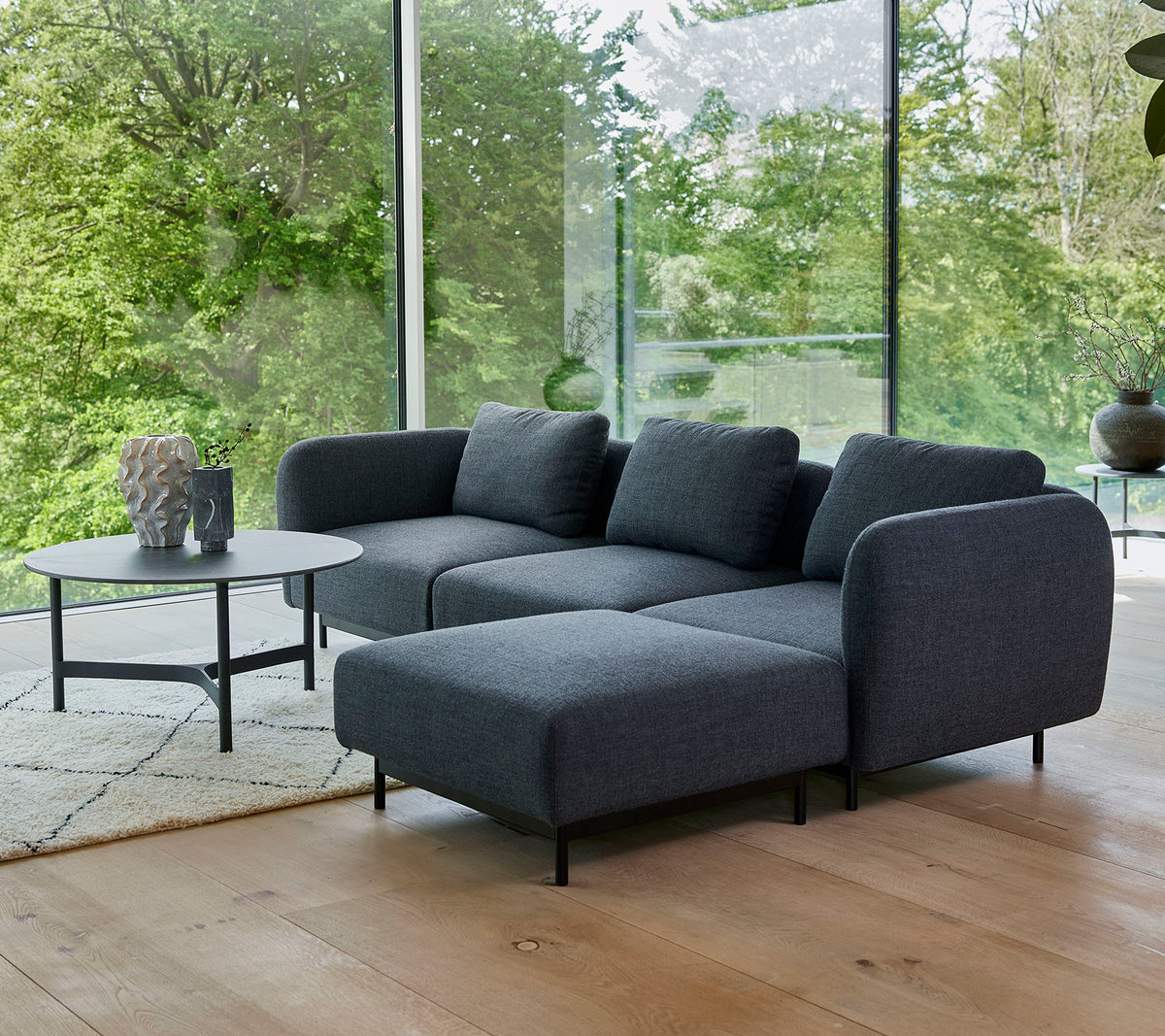 un canapé sectionnel moderne gris avec des coussins moelleux, associé à une table basse ronde élégante sur un tapis clair, entouré de verdure.