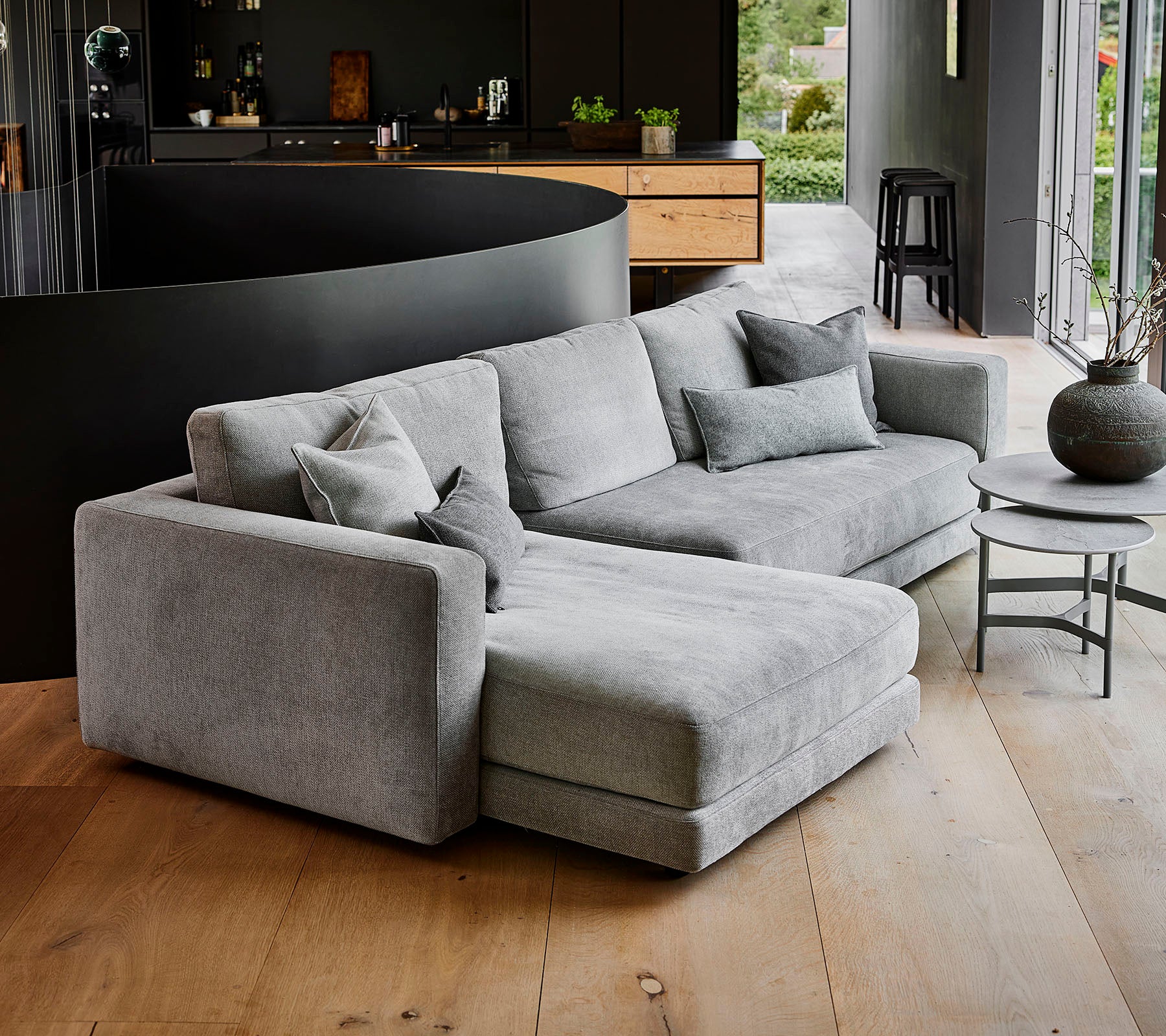 Canapé sectionnel gris moderne avec plusieurs coussins, accompagné d'une table basse ronde et d'un vase décoratif. Design simple et élégant.