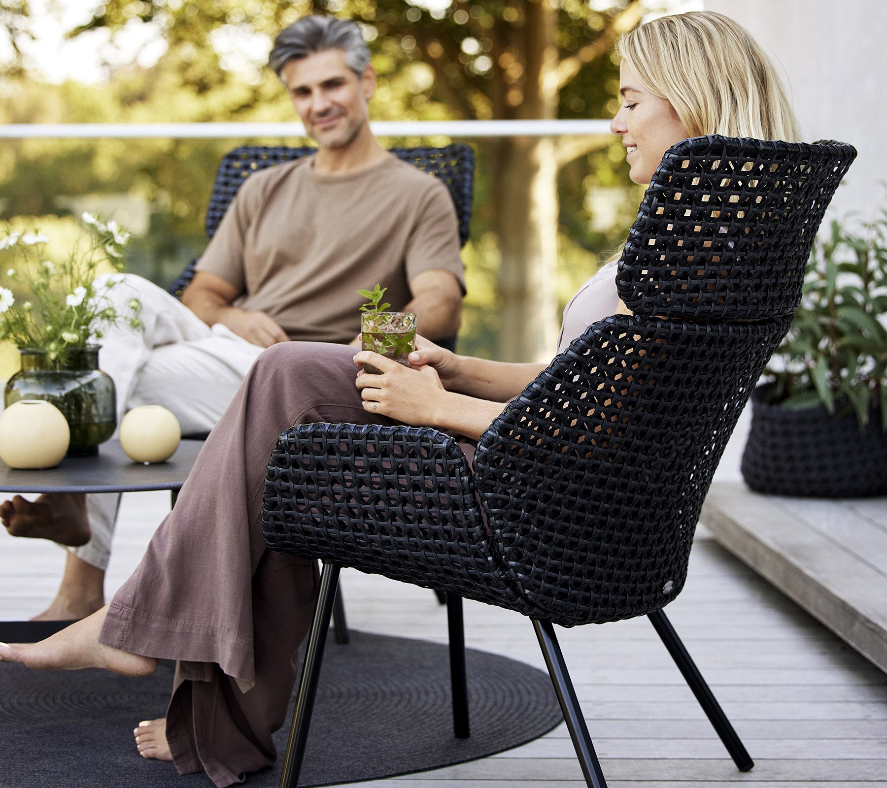 Une chaise haute et élégante en matériau tissé foncé, avec deux personnes profitant d'un moment de détente avec des plantes et de la décoration autour d'elles.