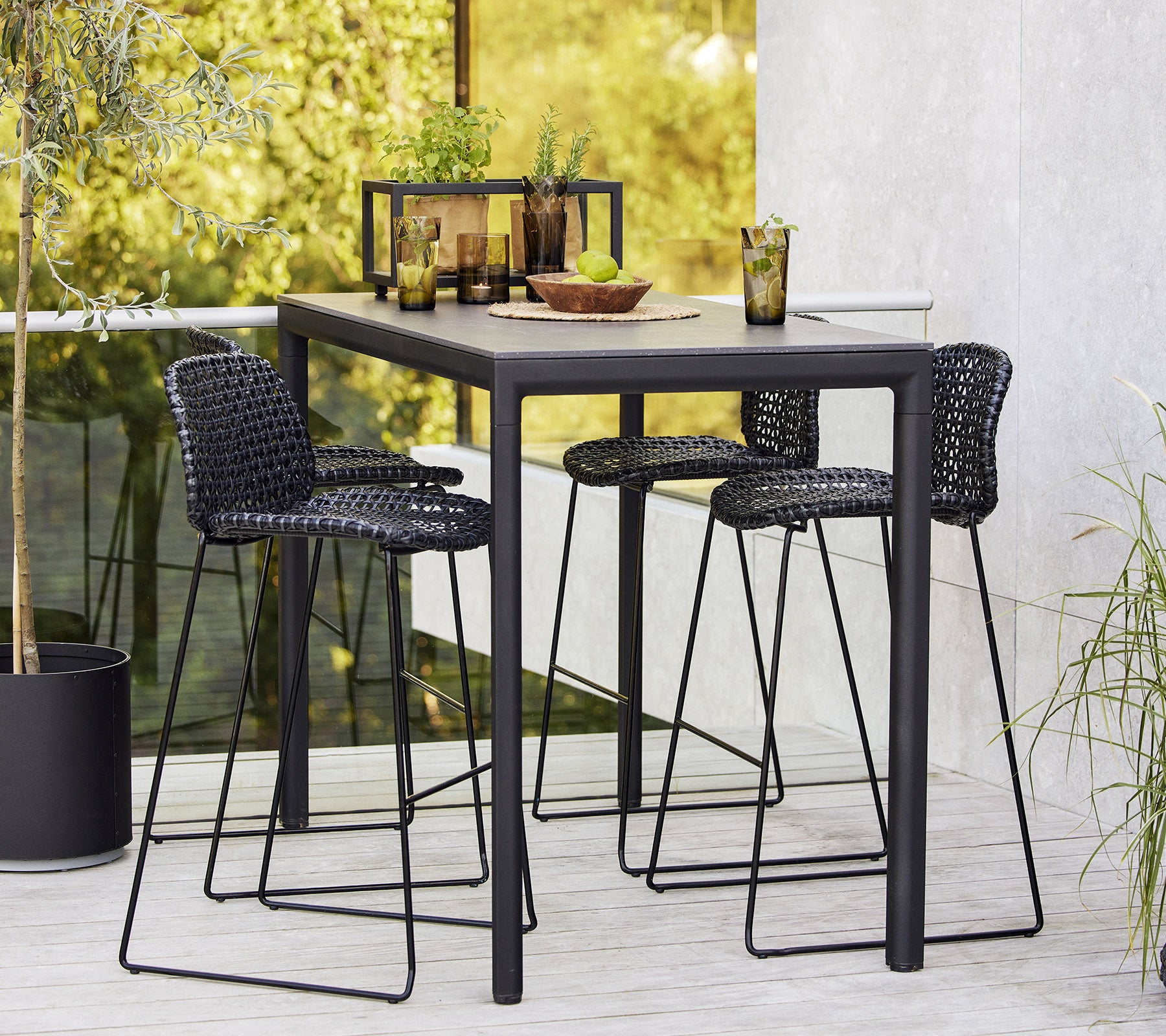 Un ensemble de table de bar élégant avec trois tabourets tressés, disposé avec de la vaisselle et des boissons, entouré de verdure.