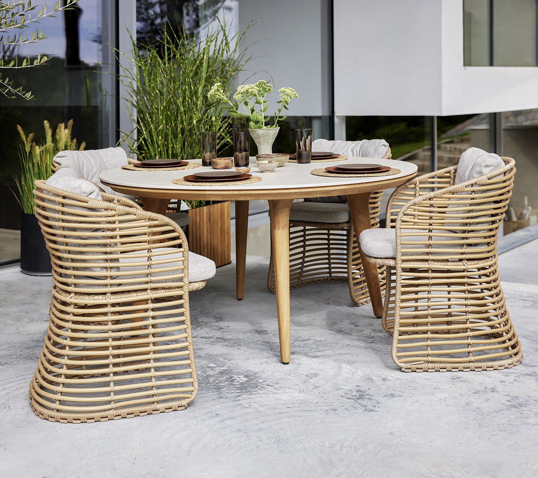 Une table ronde dressée avec quatre chaises en osier, dotées de coussins, entourée de verdure pour une salle à manger élégante.