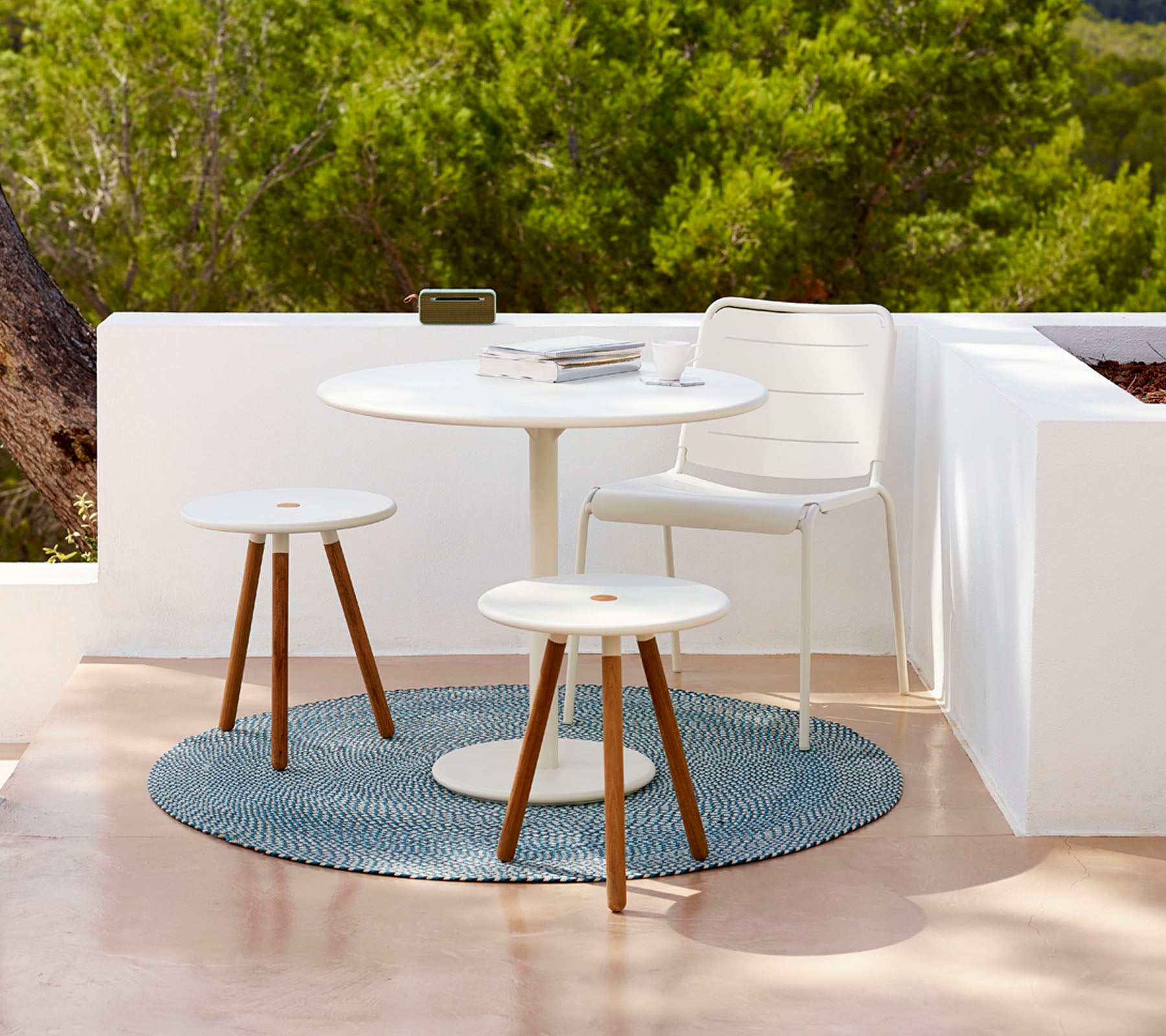 Ensemble de table d'extérieur moderne avec une table ronde, deux tabourets et un tapis texturé, entouré de verdure. Idéal pour se détendre.