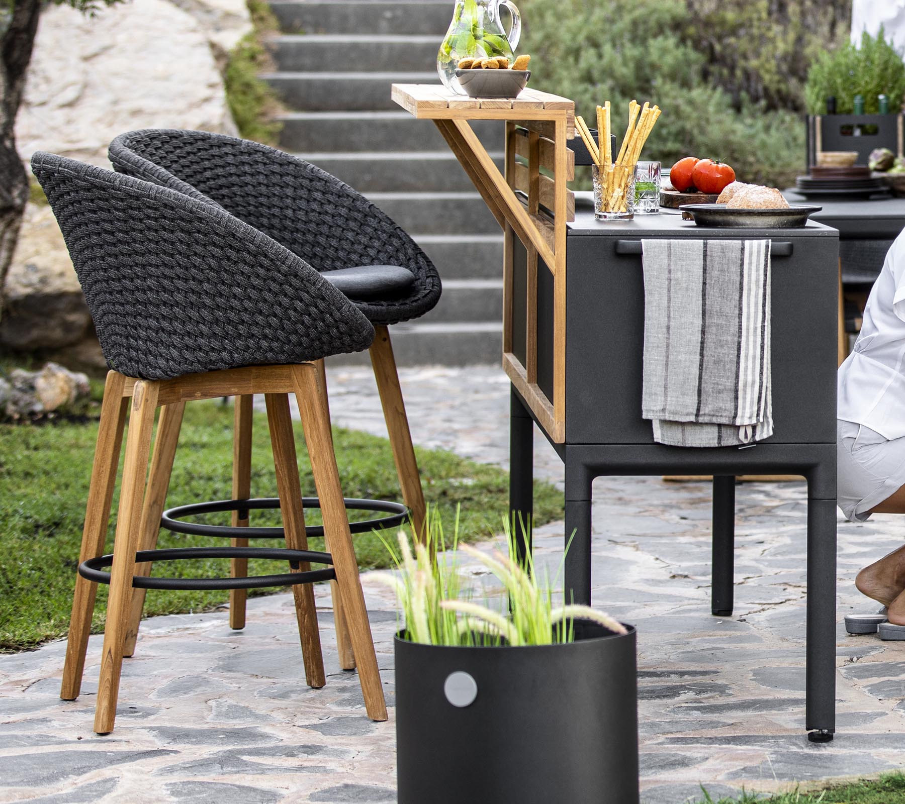 Espace extérieur élégant avec des chaises en rotin, un chariot bar moderne et des ingrédients frais disposés sur un plan de travail, agrémenté de plantes décoratives.